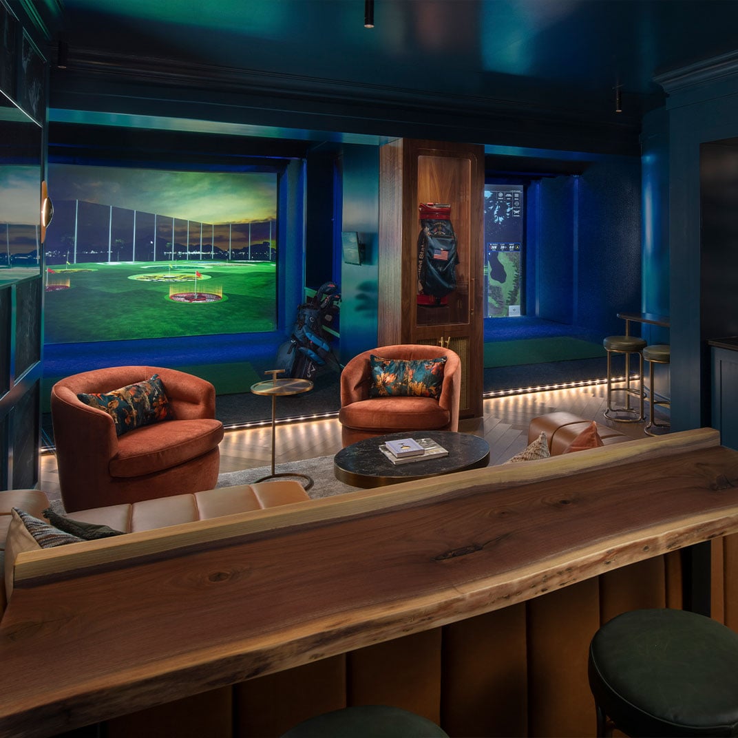 Ambient lighting bathes plush terracotta armchairs and a sleek wooden bar while golf simulators create an upscale clubhouse atmosphere in this entertainment haven.