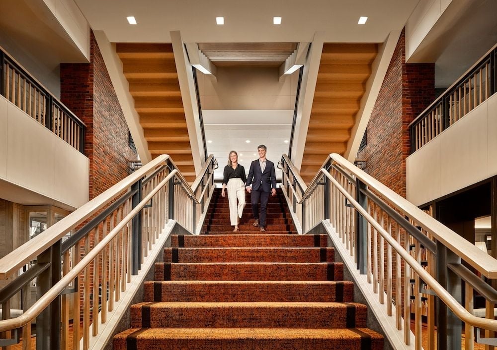 Warm brick walls and polished wood create an inviting grand staircase where couples descend hand-in-hand, illuminated by soft recessed lighting overhead.