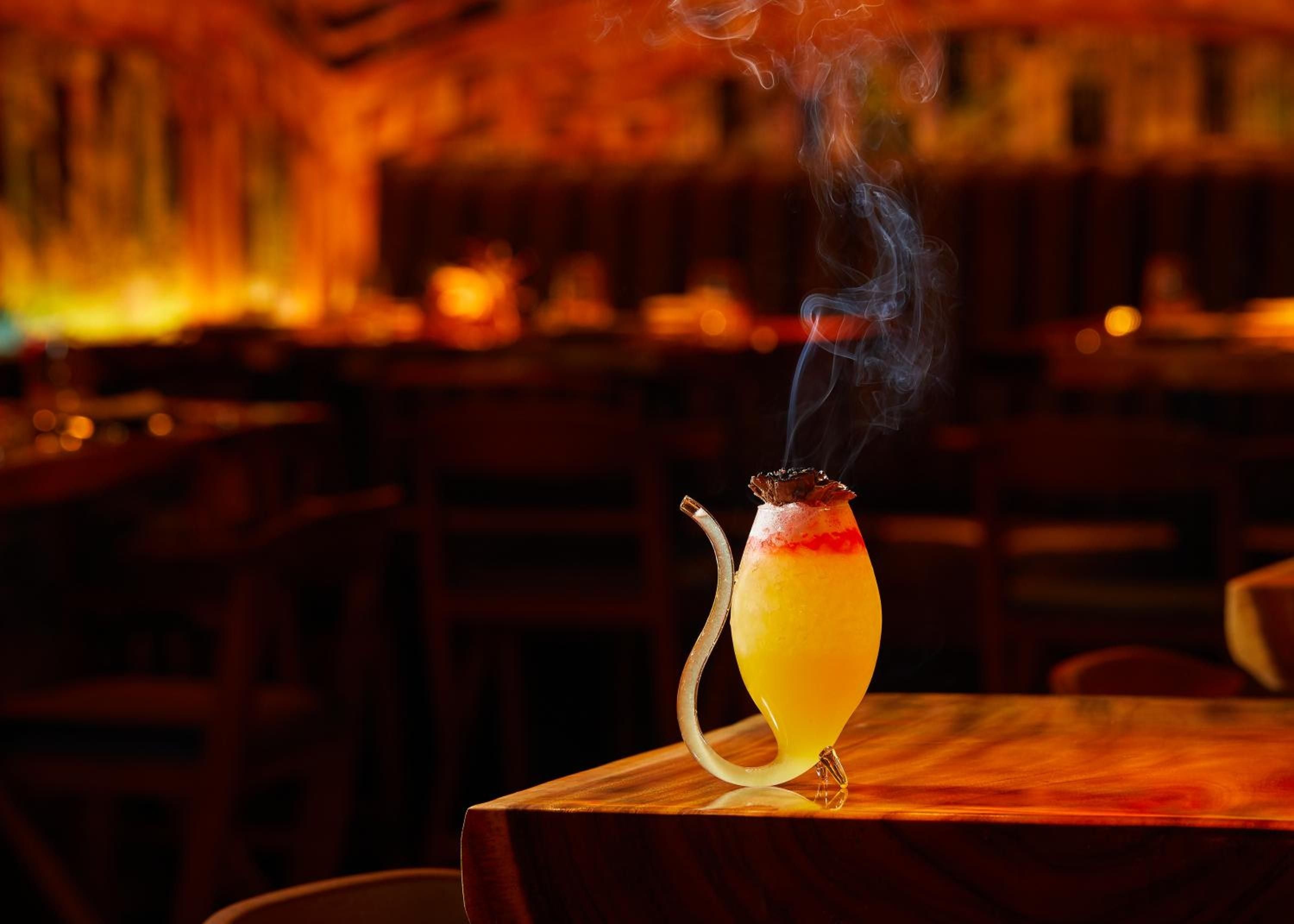 Theatrical smoke rises from a meticulously crafted cocktail at the hotel's intimate bar, where golden ambient lighting transforms evening drinks into artful experiences.