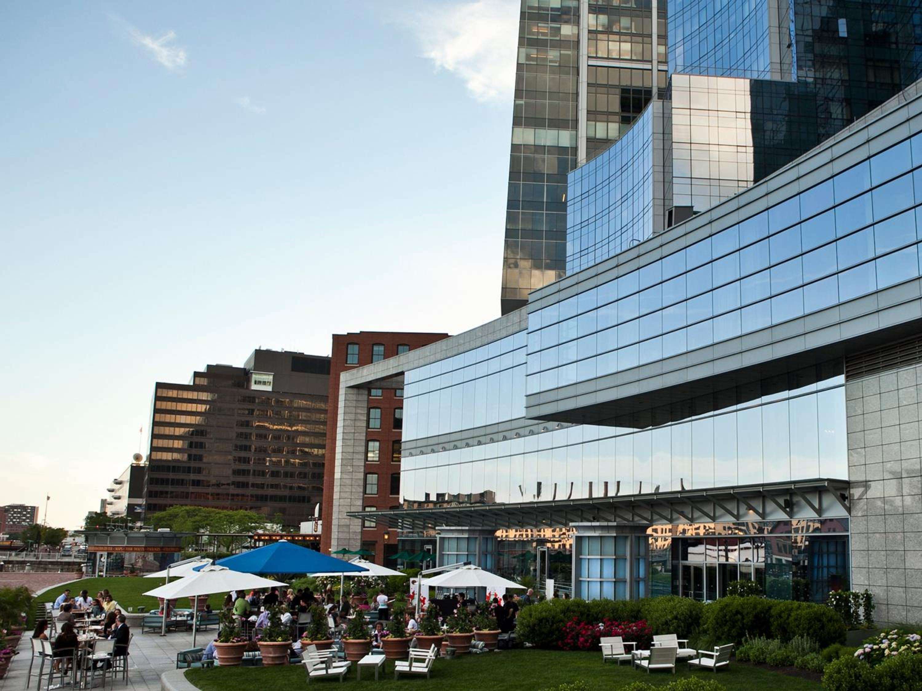 Guests gather beneath colorful umbrellas on the hotel's landscaped rooftop terrace, where modern architecture meets urban garden sanctuary against Boston's gleaming skyline.
