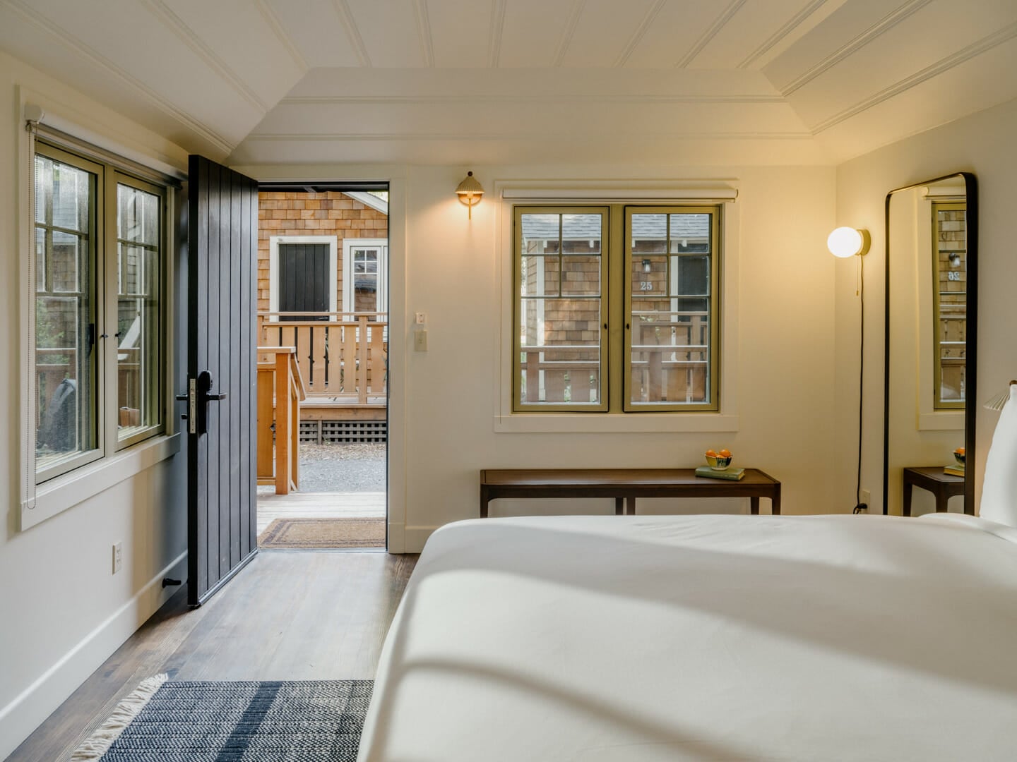 Crisp white linens and warm wood tones create a serene retreat, where folding doors open to reveal shingled cottages and wooden decks beyond.