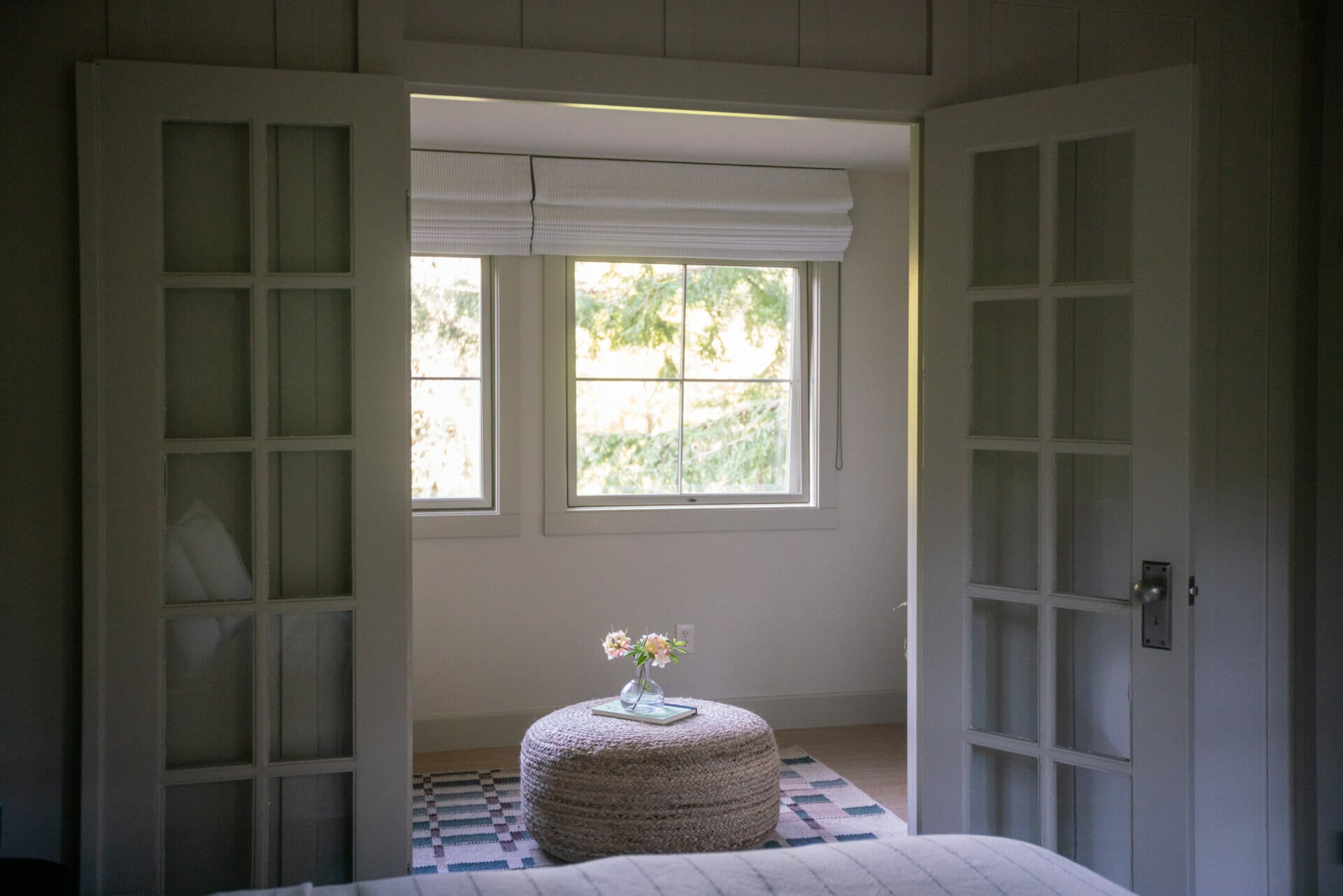 Woven rope ottoman and fresh blooms create an intimate reading nook, where geometric rugs soften polished floors and natural light filters through custom Roman shades.