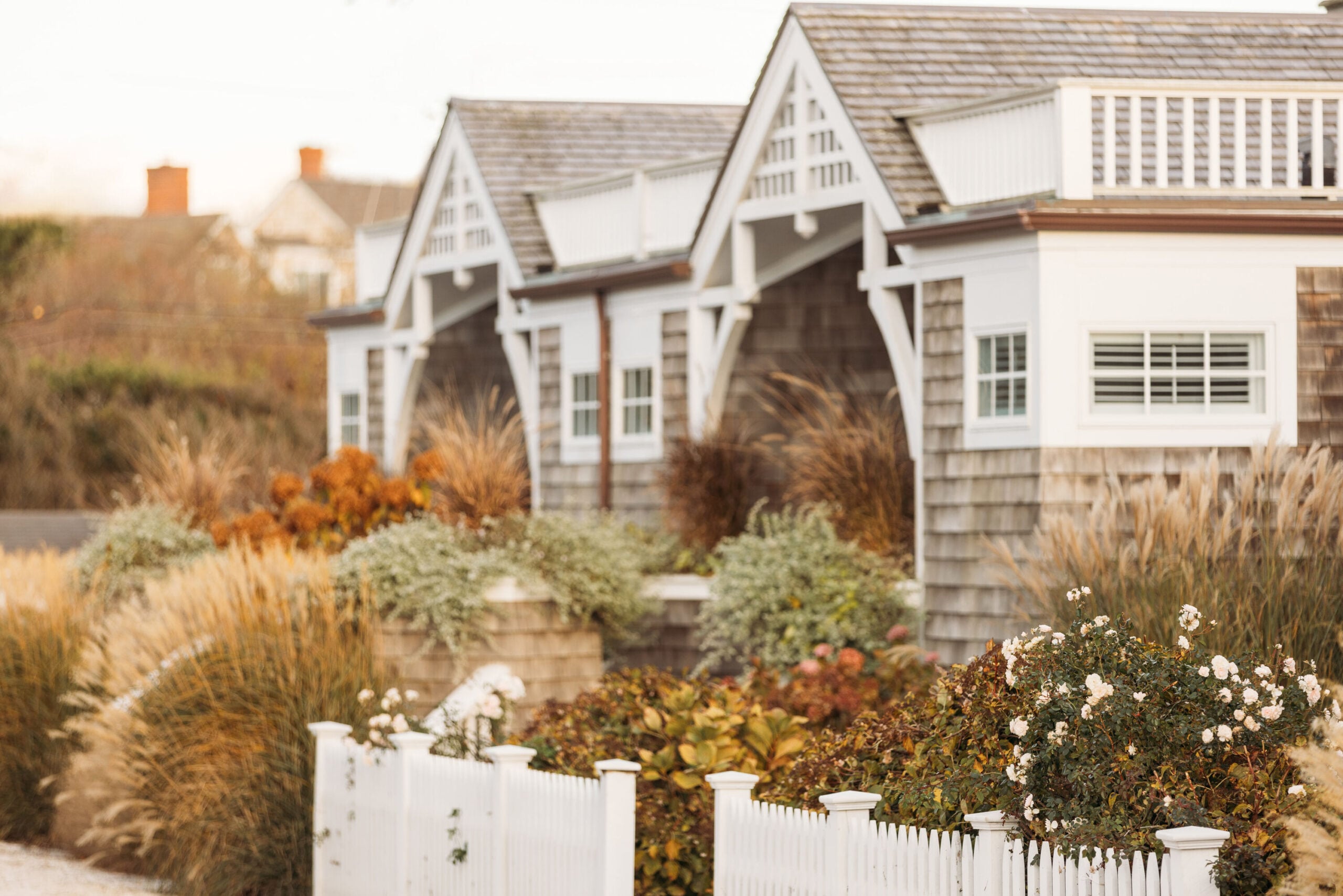Weathered shingle cottages nestle behind white picket fences and autumn ornamental grasses, where golden hour light catches the intimate charm of seaside New England architecture.