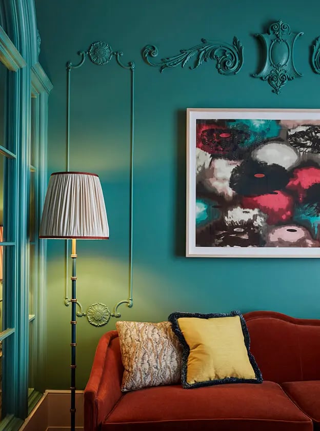 Jewel-toned walls embrace a burgundy velvet sofa where abstract art mingles with ornate moldings, creating an unexpectedly bold sanctuary of old-world elegance and contemporary flair.