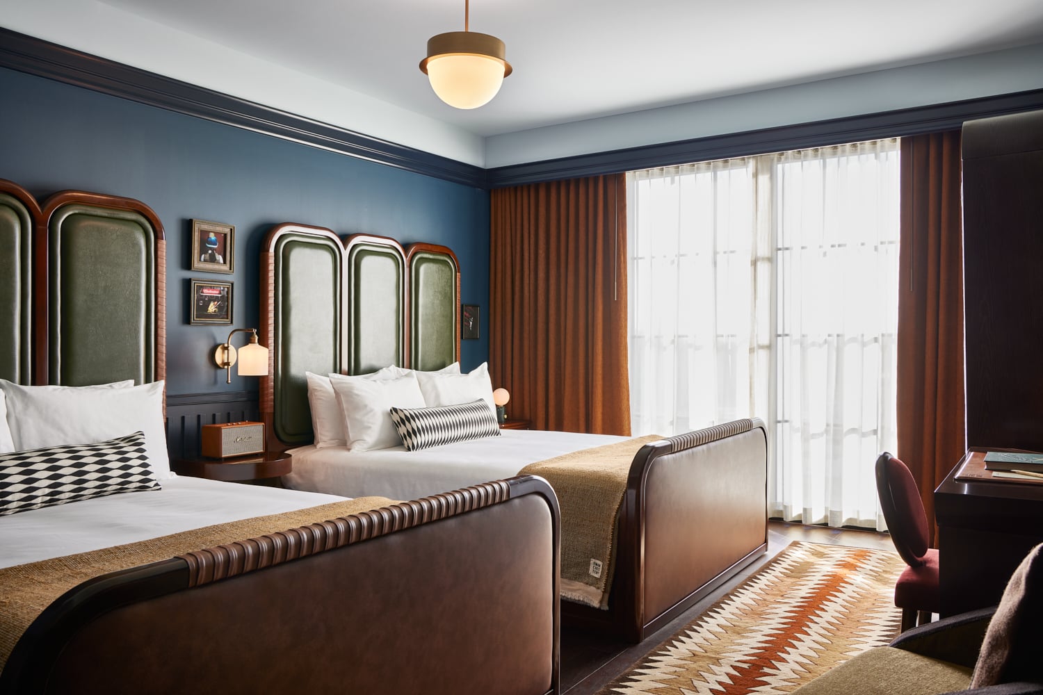Curved headboards in sage velvet frame twin beds beneath moody blue walls, while burnt orange curtains filter Manhattan light through geometric patterns.