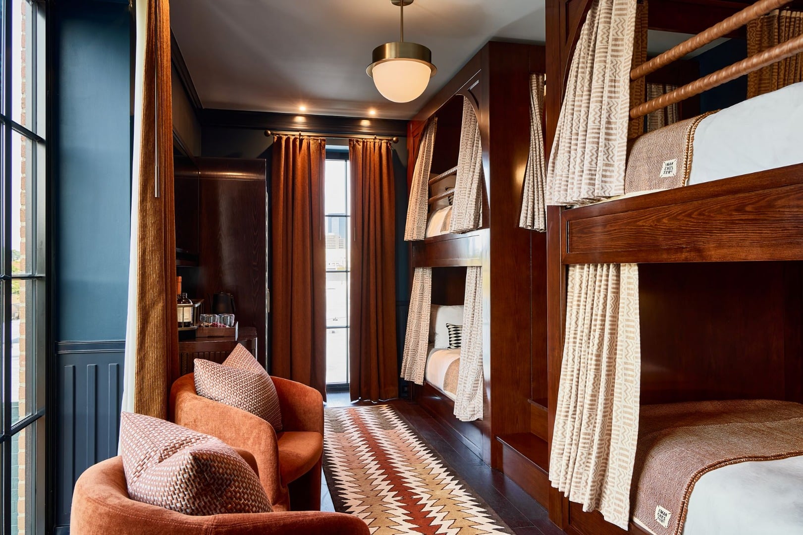 Rich walnut bunk beds with geometric curtains create an intimate sanctuary, where burnt orange velvet chairs and navy walls evoke a sophisticated mountain lodge atmosphere.