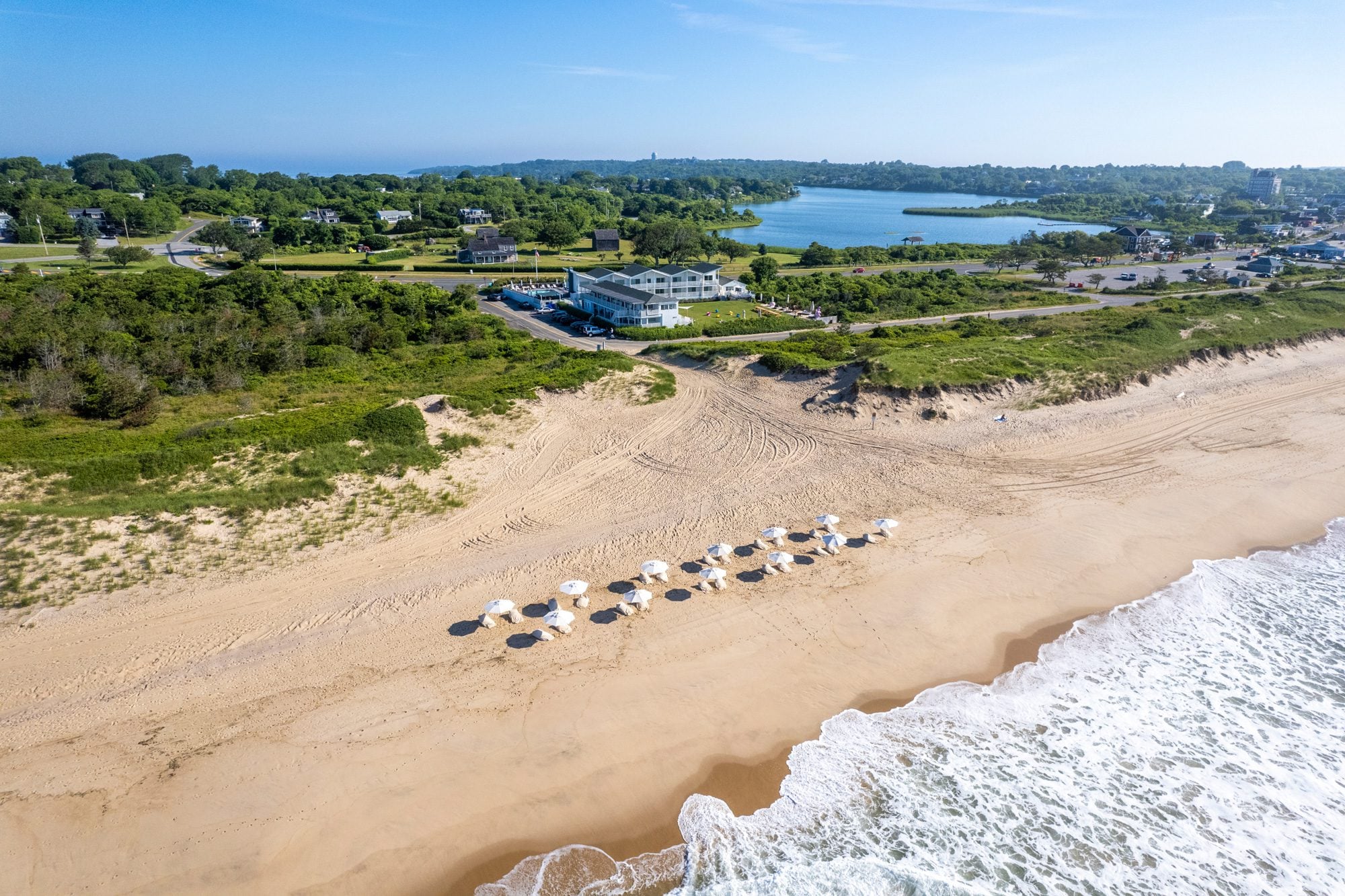 White umbrellas dot pristine sand while Hero Beach Club's sleek architecture commands the dunes, backdropped by tranquil ponds and rolling green coastline.
