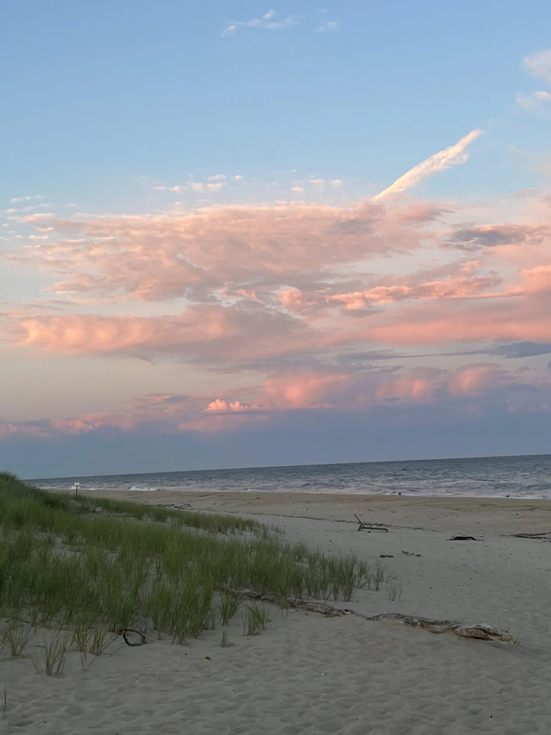 Pink-kissed clouds drift over pristine dunes where beach grass sways in the evening breeze, creating an ethereal backdrop for contemplative seaside moments.