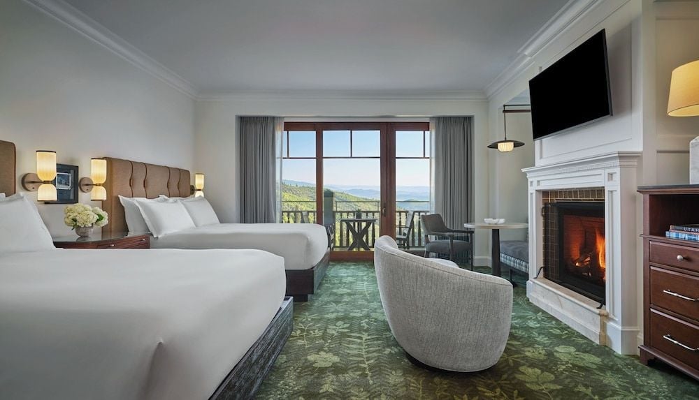 Twin beds frame a crackling fireplace while mountain vistas stretch beyond French doors, creating an intimate alpine retreat with refined rustic elegance.