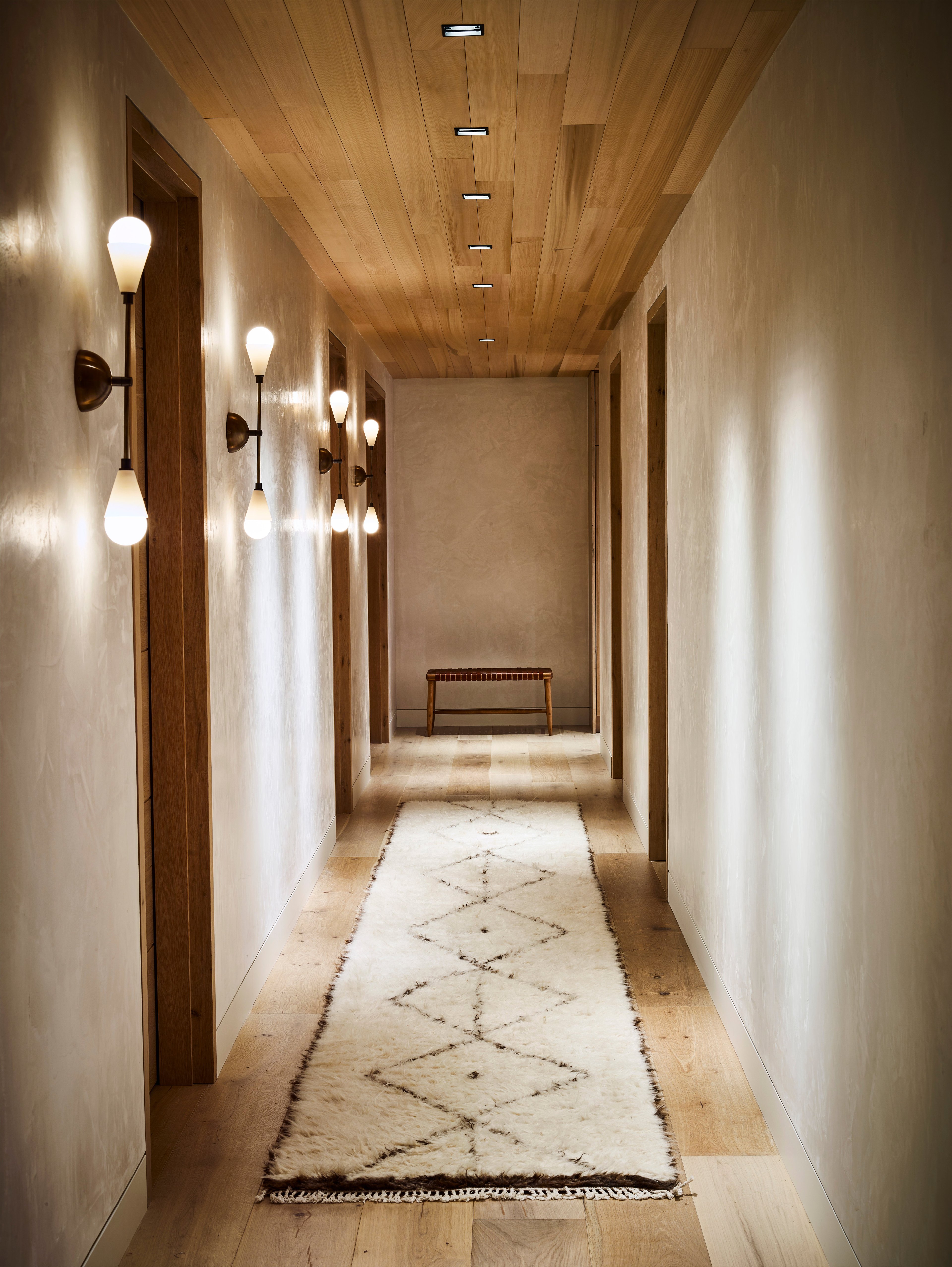 Warm wood-planked ceilings and sculptural sconces create an intimate gallery-like corridor, where handwoven rugs guide guests through softly illuminated limestone passages.