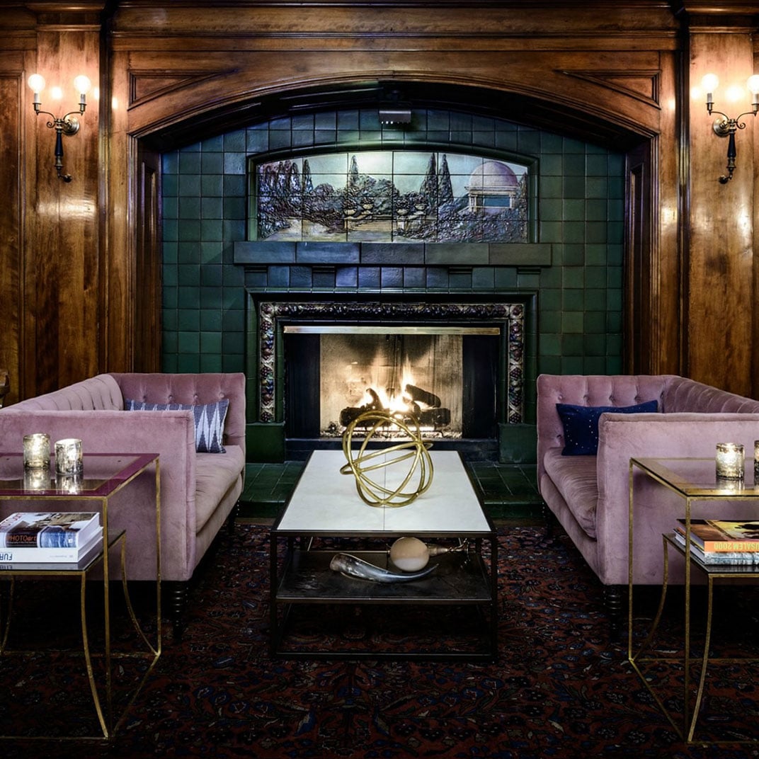 Velvet armchairs in dusty rose face a crackling fireplace beneath hand-painted tiles, where warm sconces cast golden light across rich wood paneling.