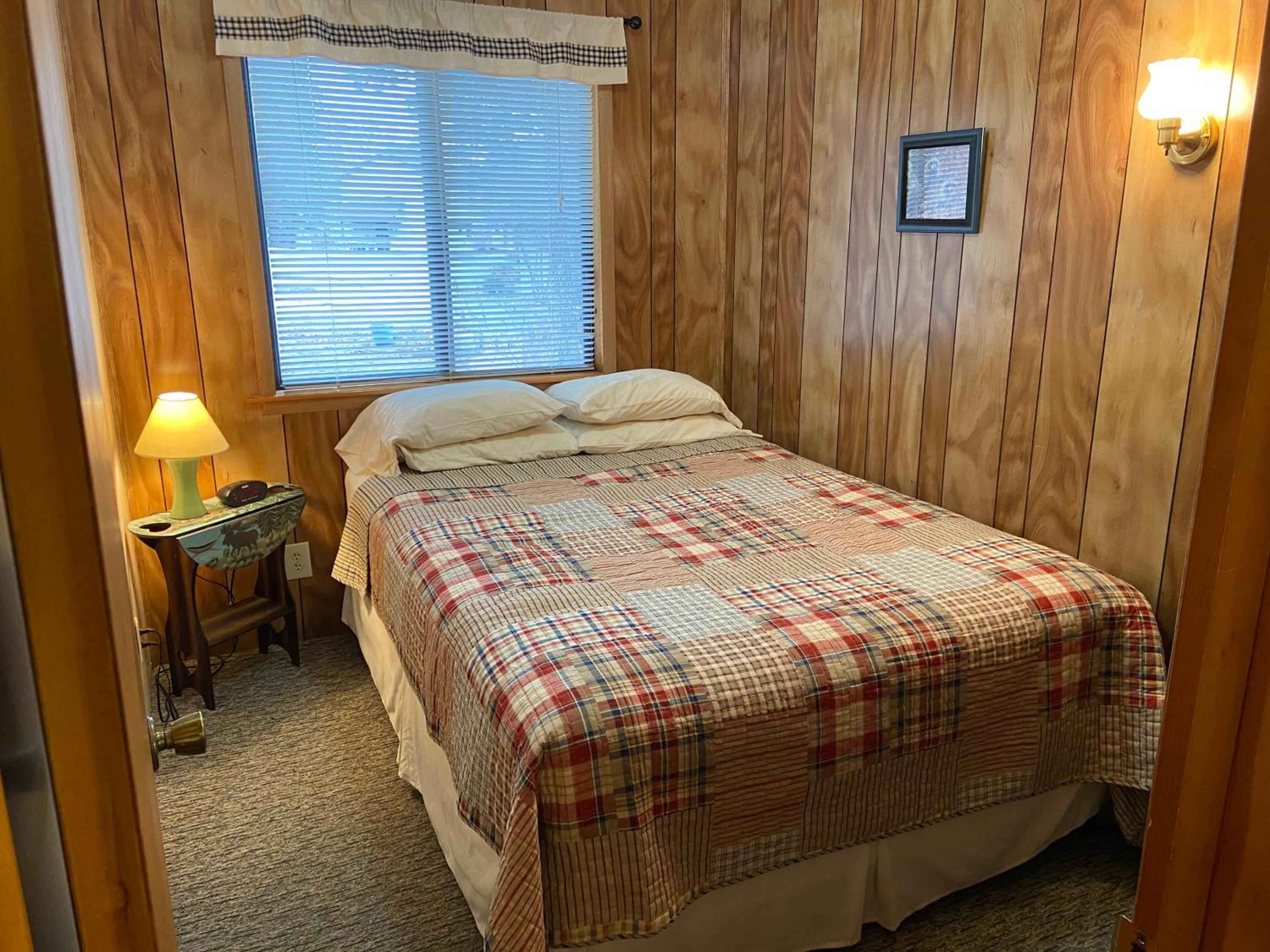 Knotty pine walls and plaid quilts create rustic charm in cozy guest rooms, where warm lamplight and blue blinds frame peaceful woodland views.