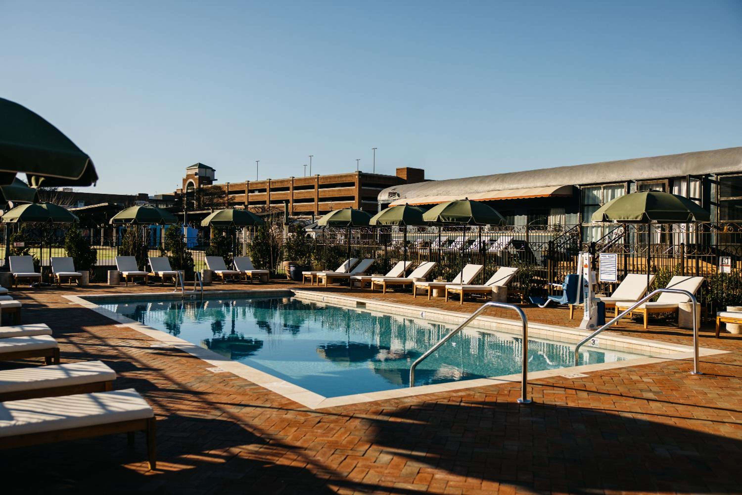 Afternoon light dances across brick pavers surrounding the crystal-clear pool, where sleek loungers invite languid hours beneath green umbrellas and open skies.