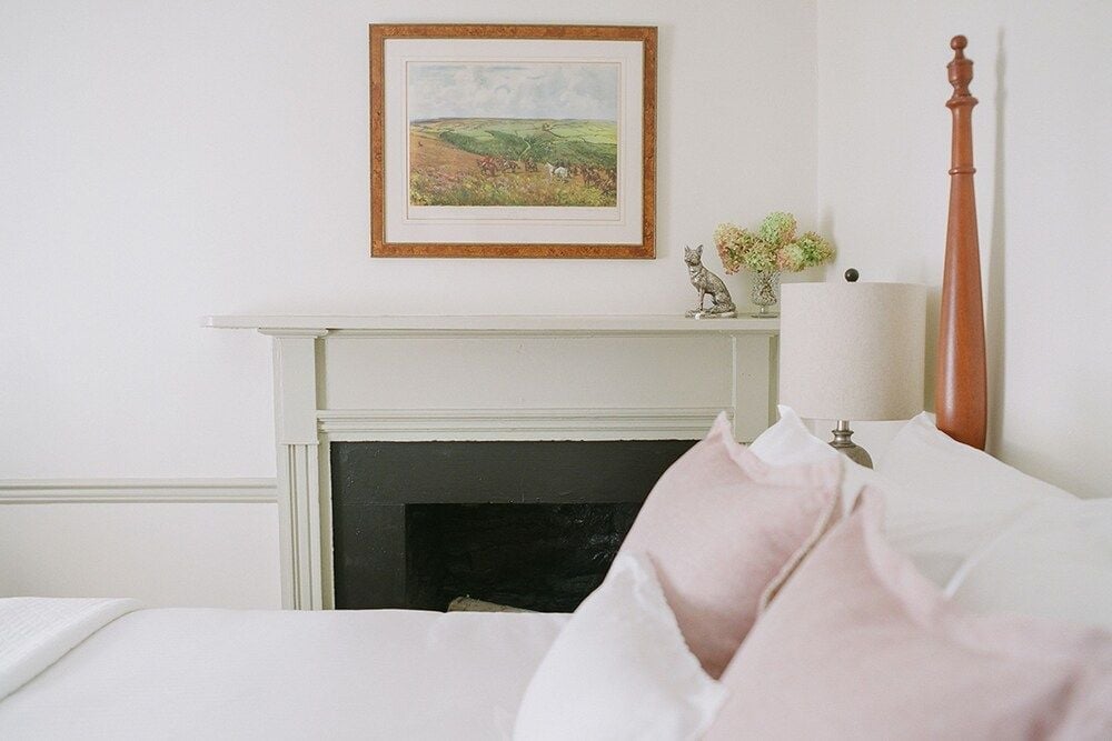 Soft morning light filters across crisp white linens and blush pillows, while a pastoral landscape painting anchors this serene guest room's classic charm.