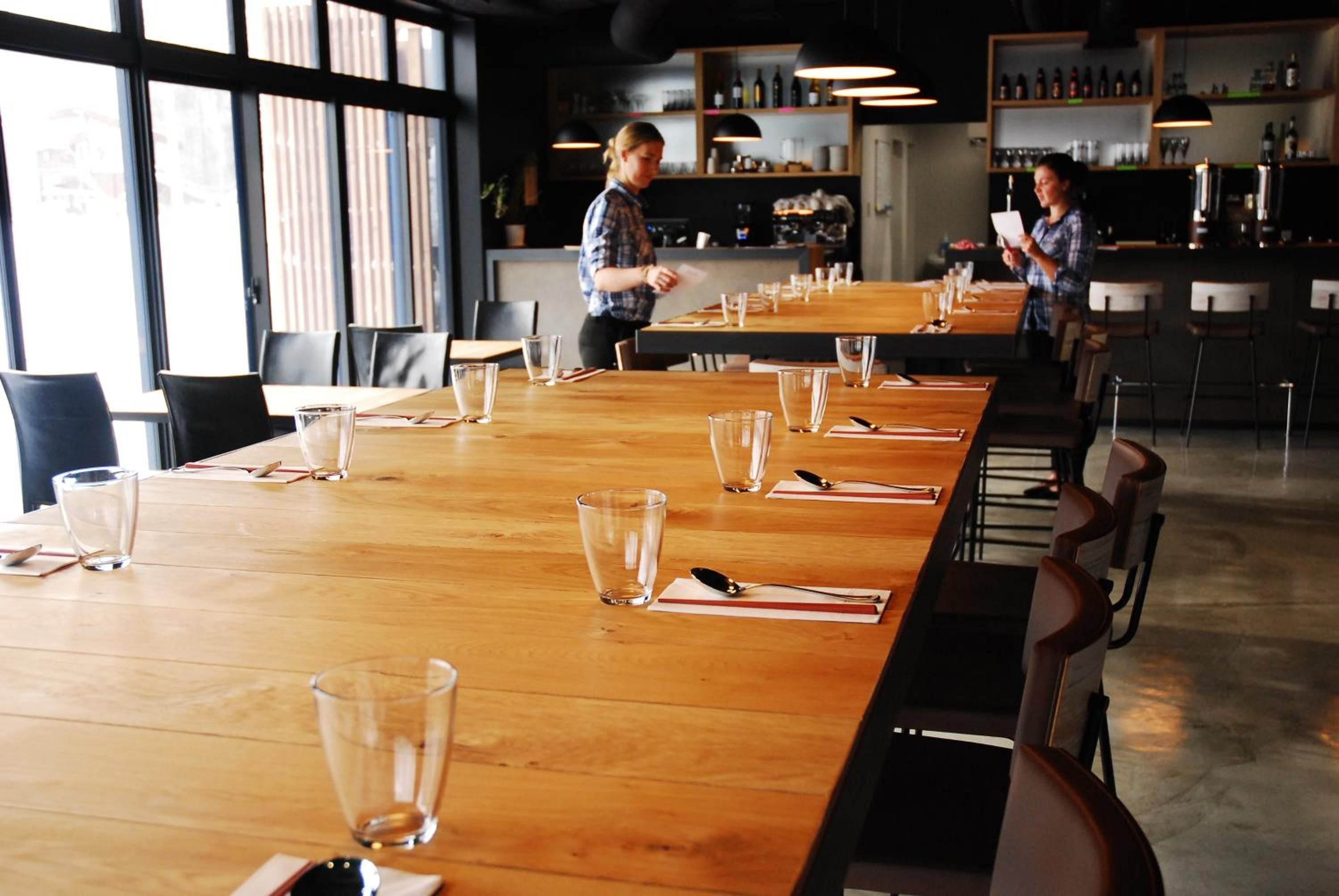 Communal wooden tables set with simple glassware create an intimate dining atmosphere where servers prepare for evening service beneath warm pendant lighting.