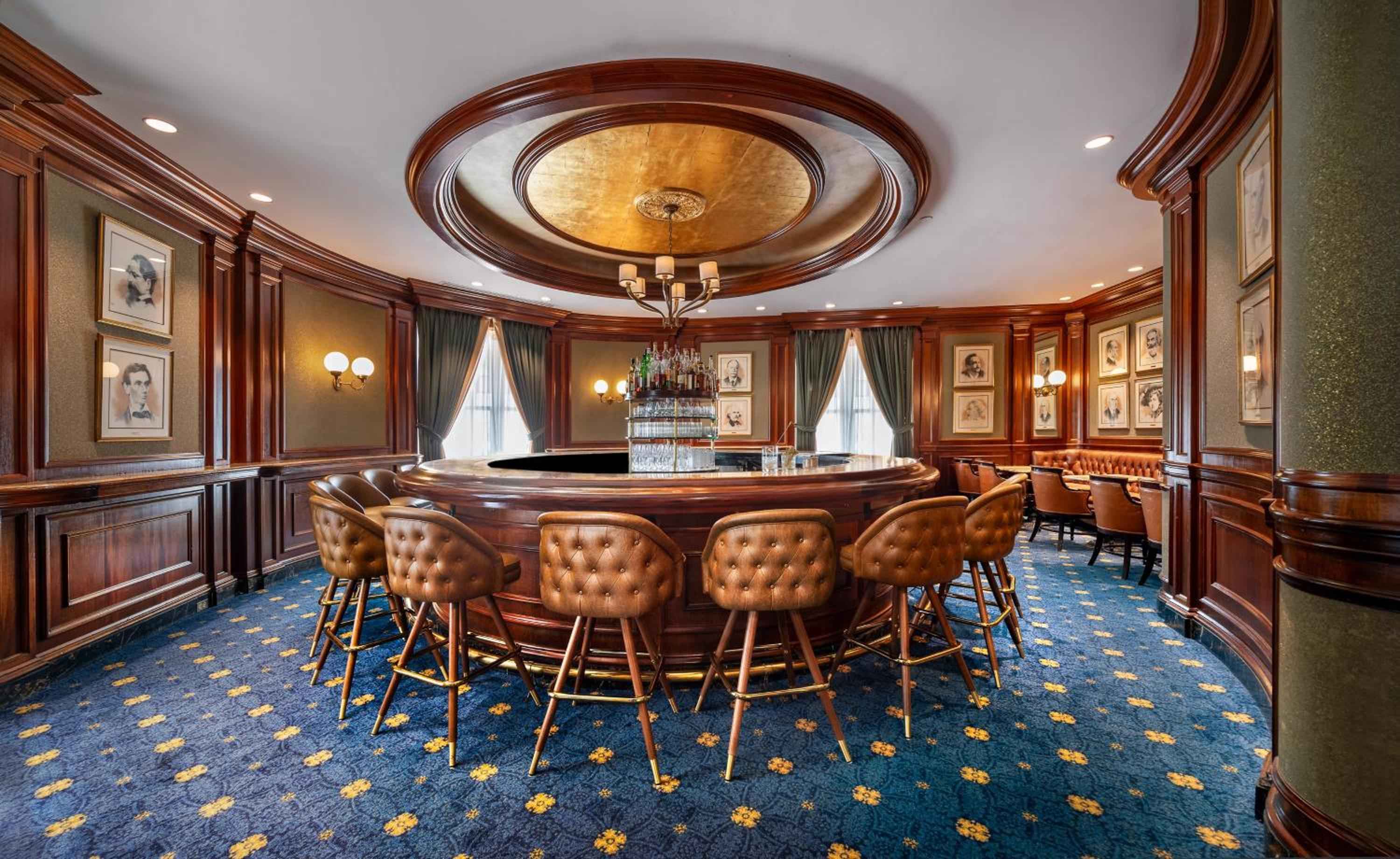 Rich mahogany paneling and tufted leather stools create an intimate gentleman's club atmosphere in this circular bar, where crystal decanters catch warm amber light beneath the gilded dome ceiling.