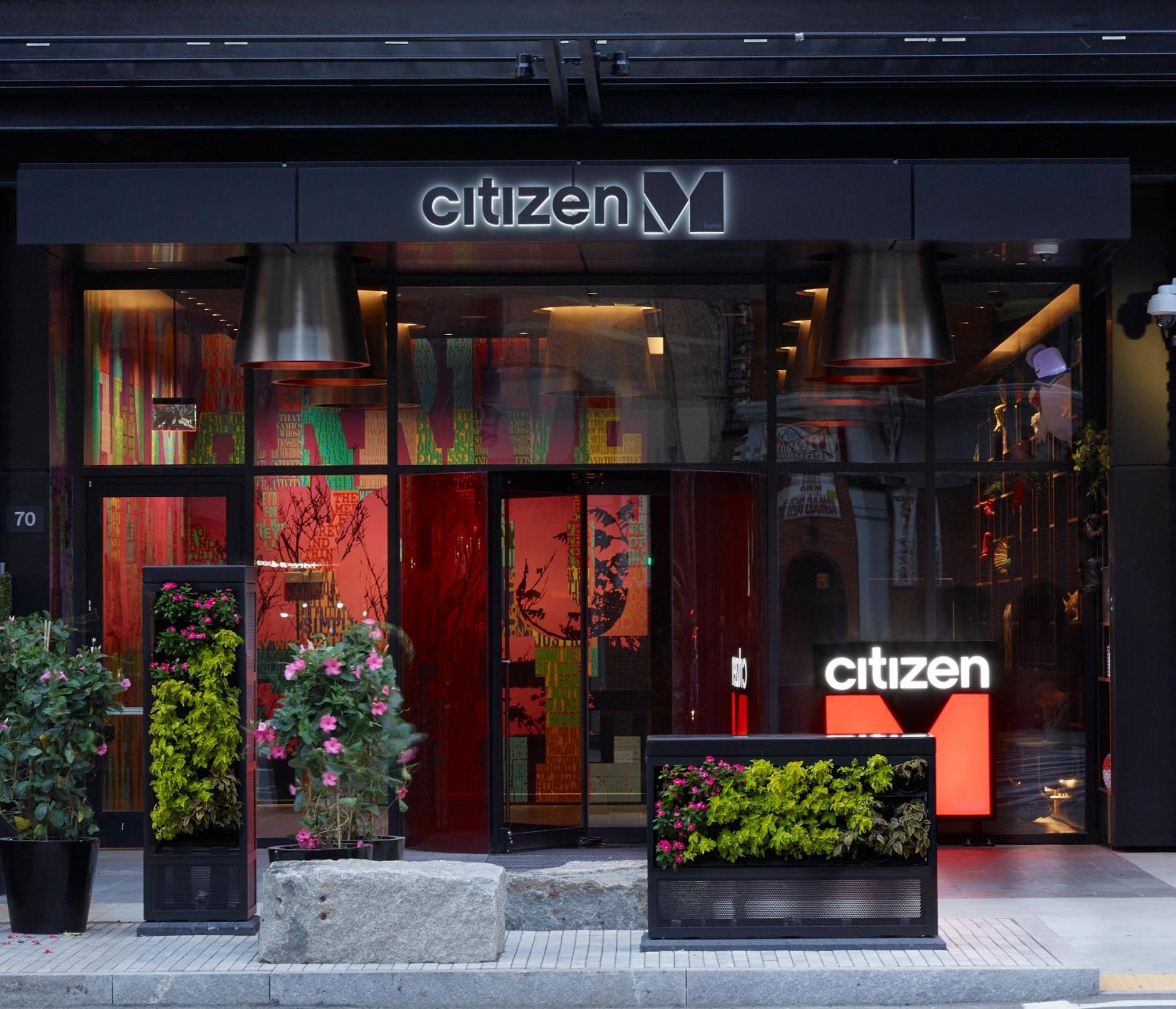 Bold orange signage and artistic window displays create an inviting street presence, while planters of vibrant blooms soften the modern facade's industrial edges.