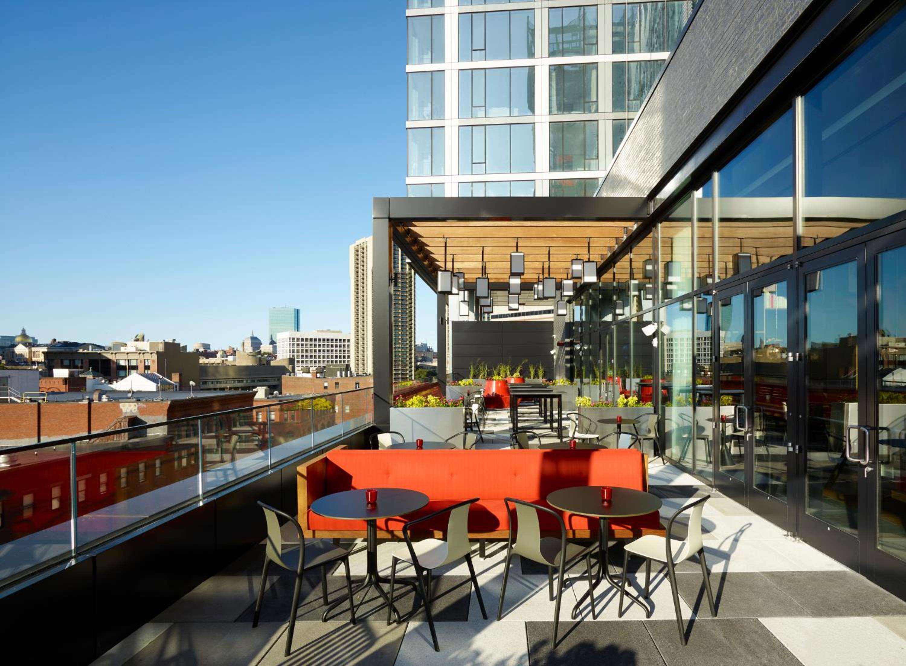 Orange banquettes and sleek bistro tables create an inviting rooftop retreat, where Boston's brick neighborhoods stretch toward gleaming downtown towers under endless blue skies.