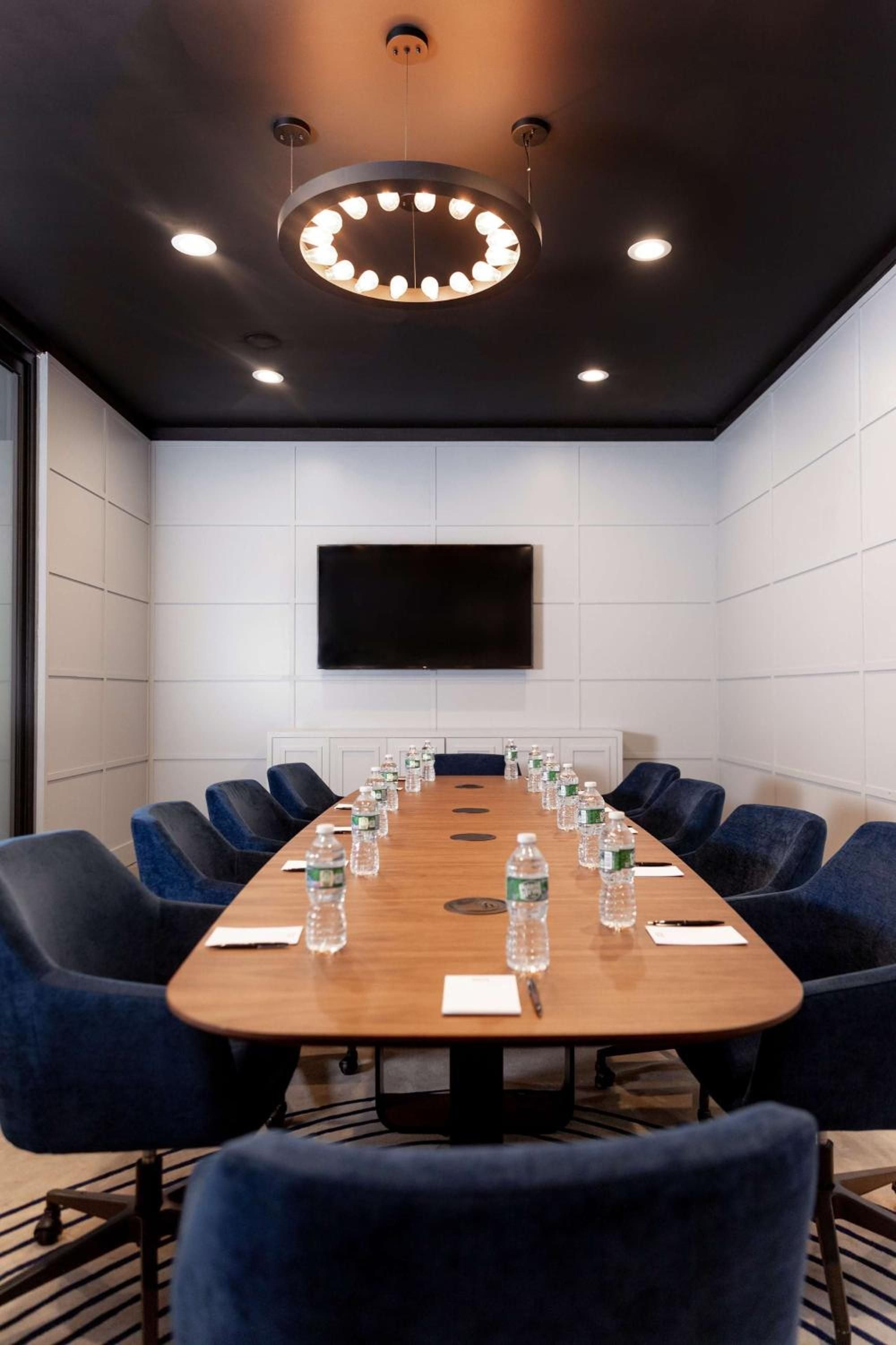 Navy velvet chairs surround a polished wood table beneath a striking circular chandelier, creating an intimate boardroom where natural light meets sophisticated design.