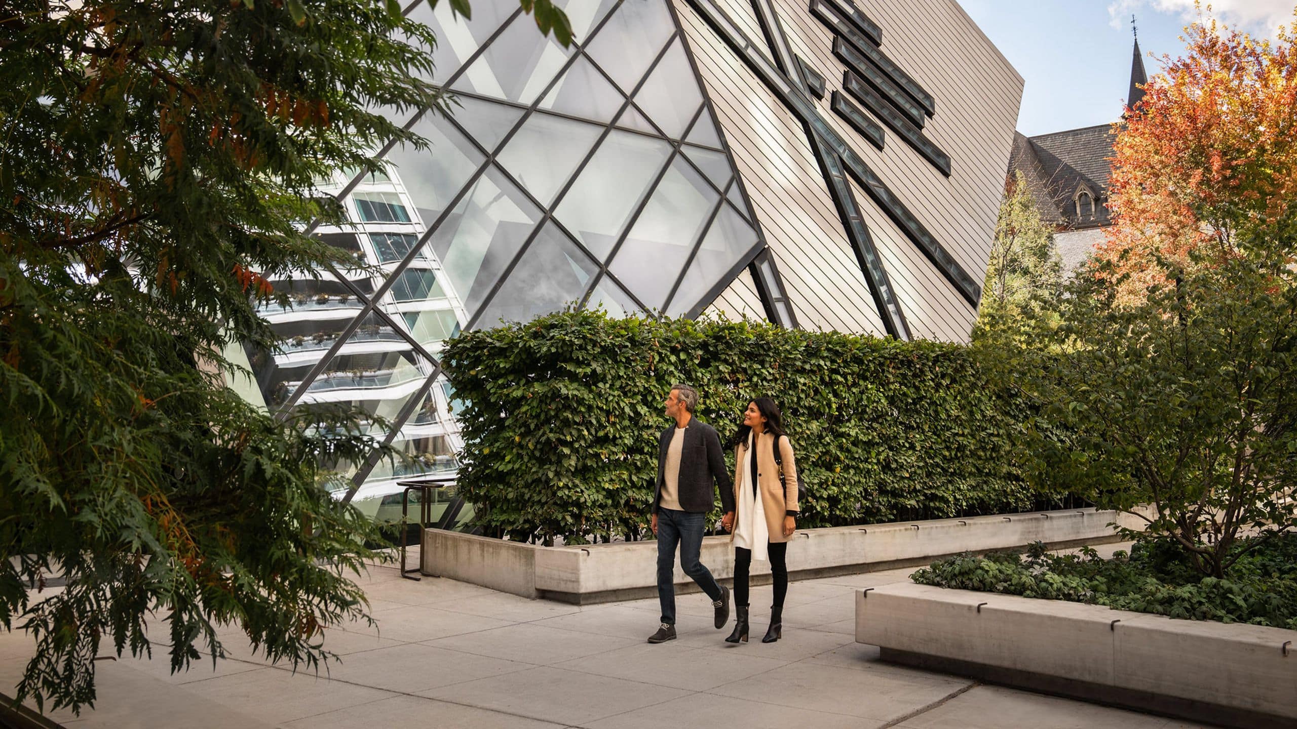 Autumn light filters through leafy branches as guests stroll past the hotel's striking angular façade, where modern geometry meets Toronto's seasonal splendor.