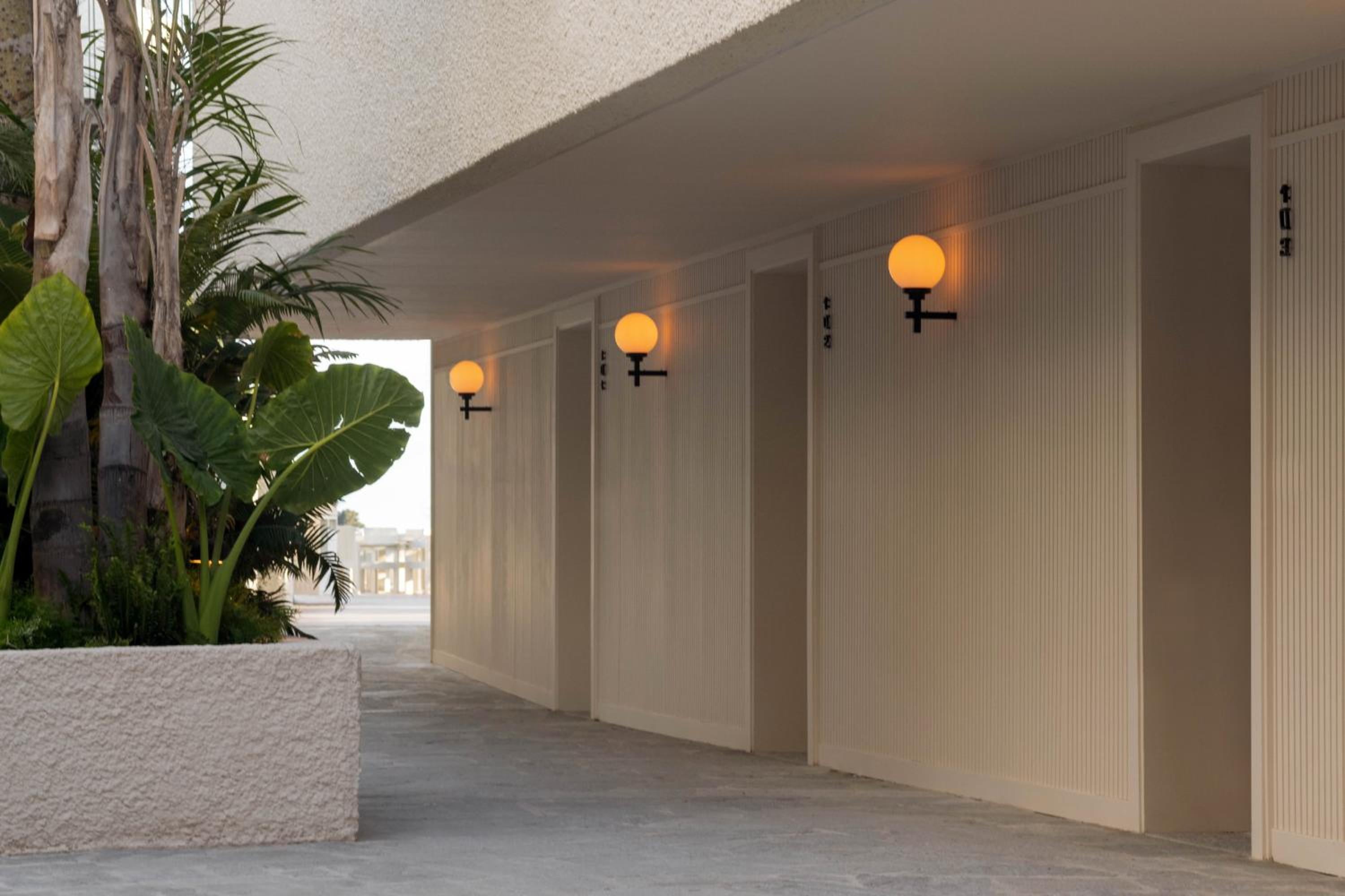 Warm globe sconces illuminate textured wood panels along the hotel's covered walkway, where tropical palms frame intimate alcoves in golden evening light.