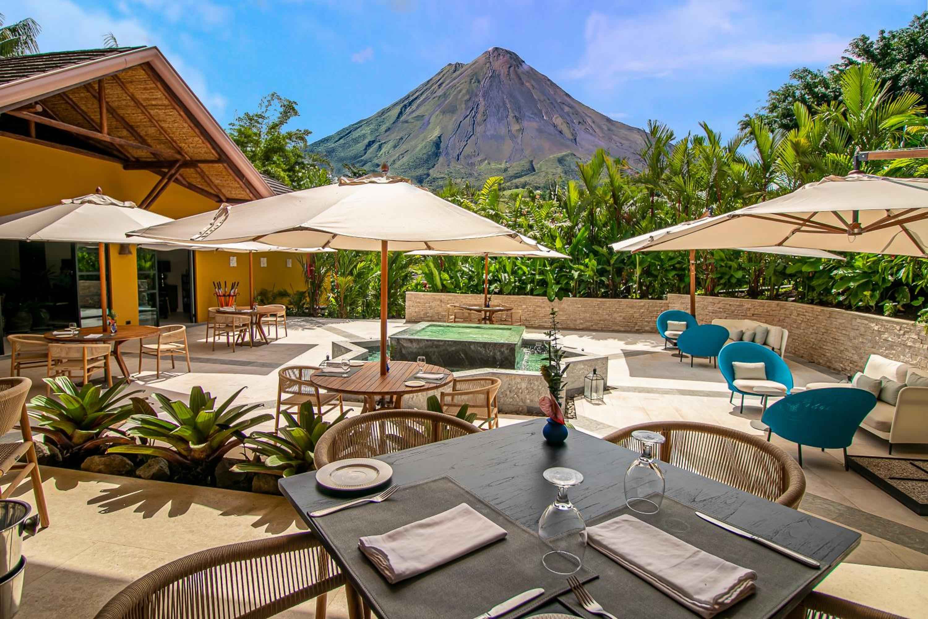 Volcanic Arenal looms majestically beyond the open-air terrace, where teak tables and woven chairs invite lingering meals beneath cream umbrellas and tropical palms.