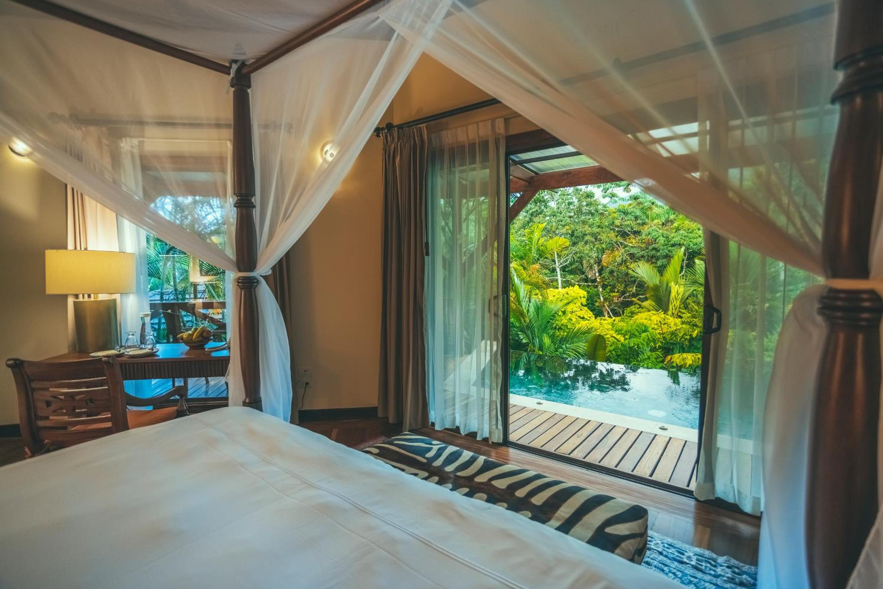 Billowing mosquito netting frames a four-poster bed that opens directly onto private infinity waters, where jungle canopy meets crystalline pools through floor-to-ceiling glass doors.