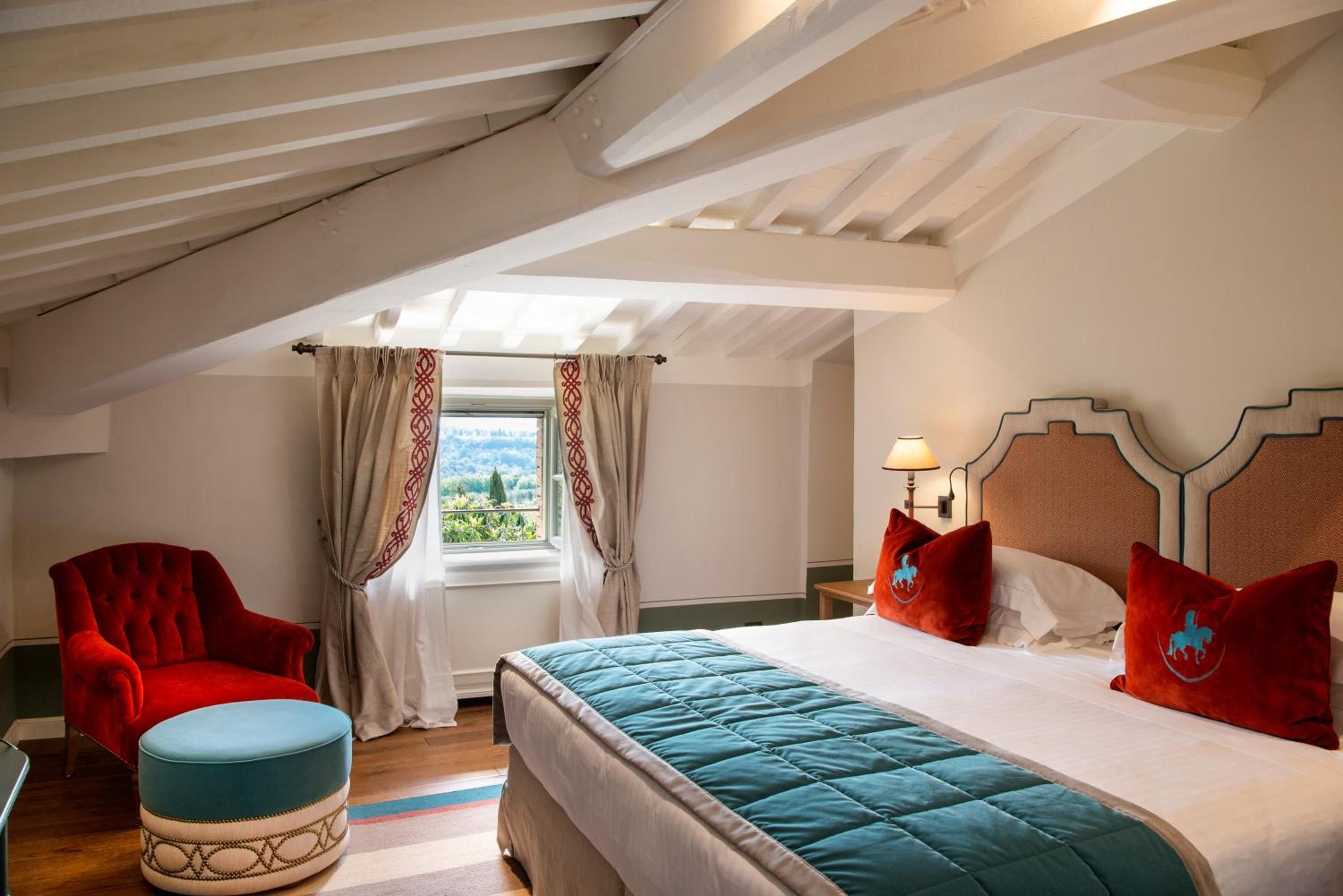 Exposed wooden beams frame this intimate attic suite, where crimson velvet chairs and turquoise accents create warmth beneath slanted ceilings overlooking Tuscan hills.