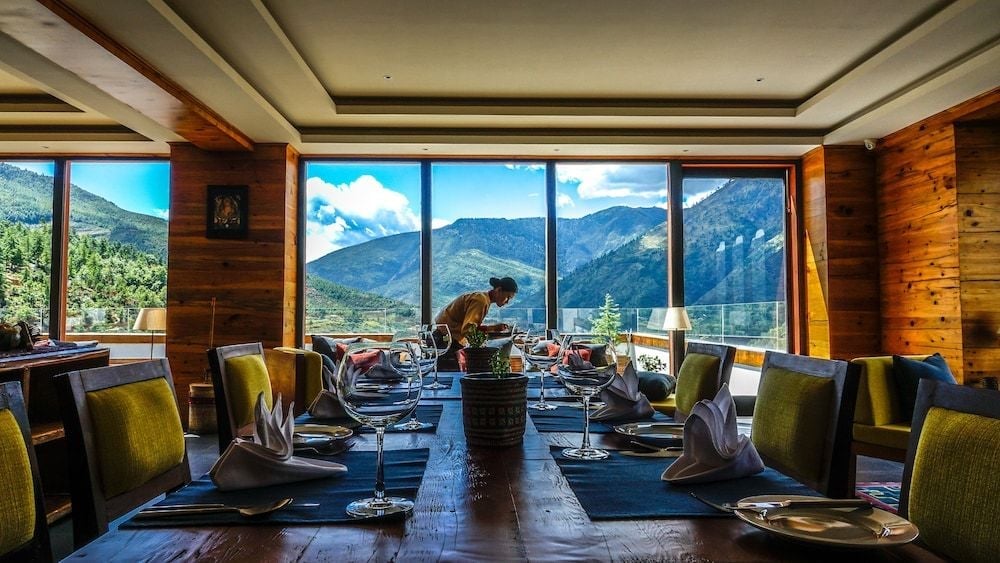 Floor-to-ceiling windows frame the dramatic Thimphu valley while a server tends to elegantly set tables in this warmly lit mountain dining room.
