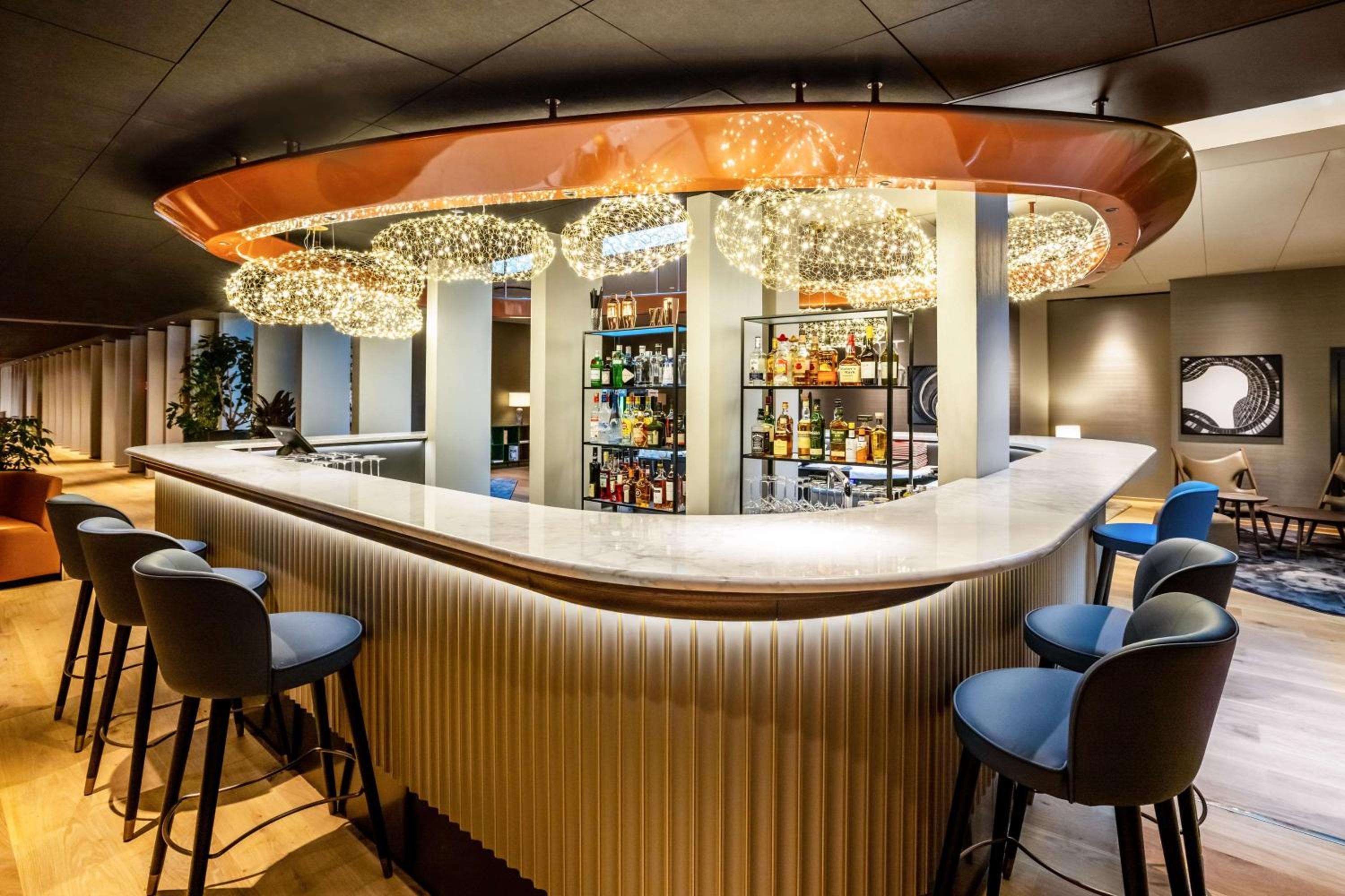 Sparkling chandeliers cast golden light over the curved marble bar, where blue velvet stools invite intimate conversations amid gleaming bottles and warm amber accents.