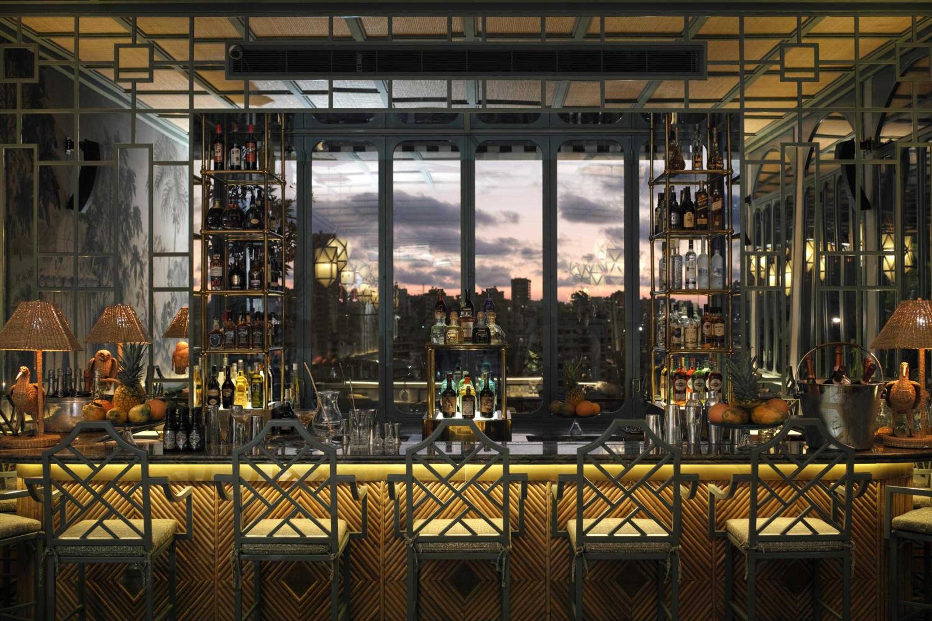Golden hour light filters through floor-to-ceiling windows as the sophisticated bar showcases premium spirits in illuminated glass towers, creating an intimate yet grand drinking sanctuary.
