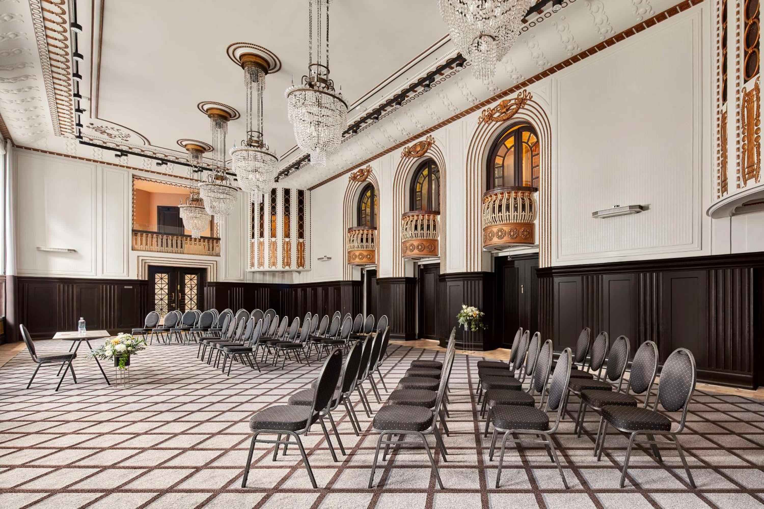 Crystal chandeliers illuminate ornate columns and dark wood wainscoting in this grand ballroom, where gilded details and checkered floors create an atmosphere of old-world elegance.