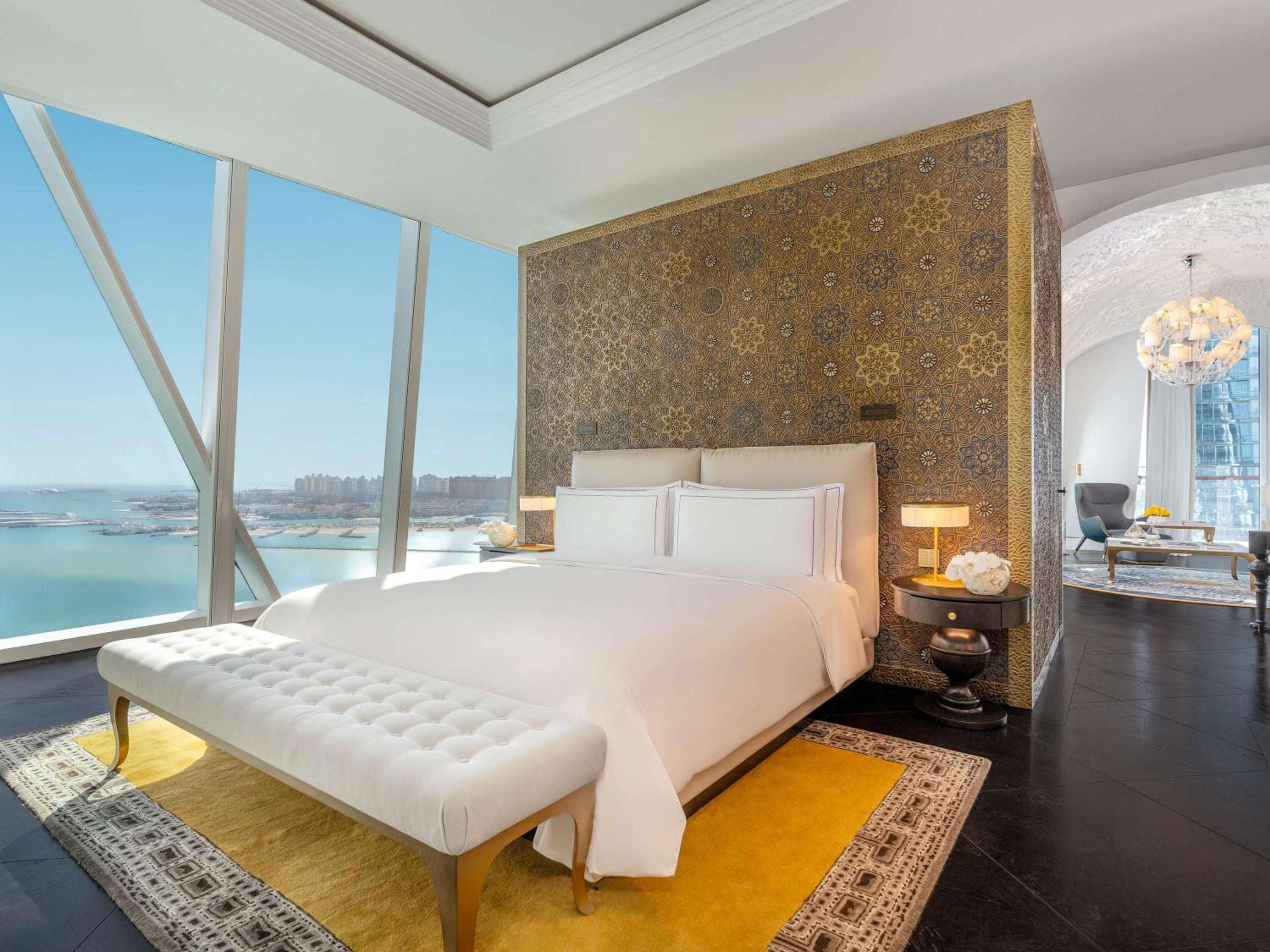 Floor-to-ceiling windows frame turquoise Arabian Gulf waters while intricate geometric wallpaper and crisp white linens create an elegant sanctuary high above Doha's coastline.
