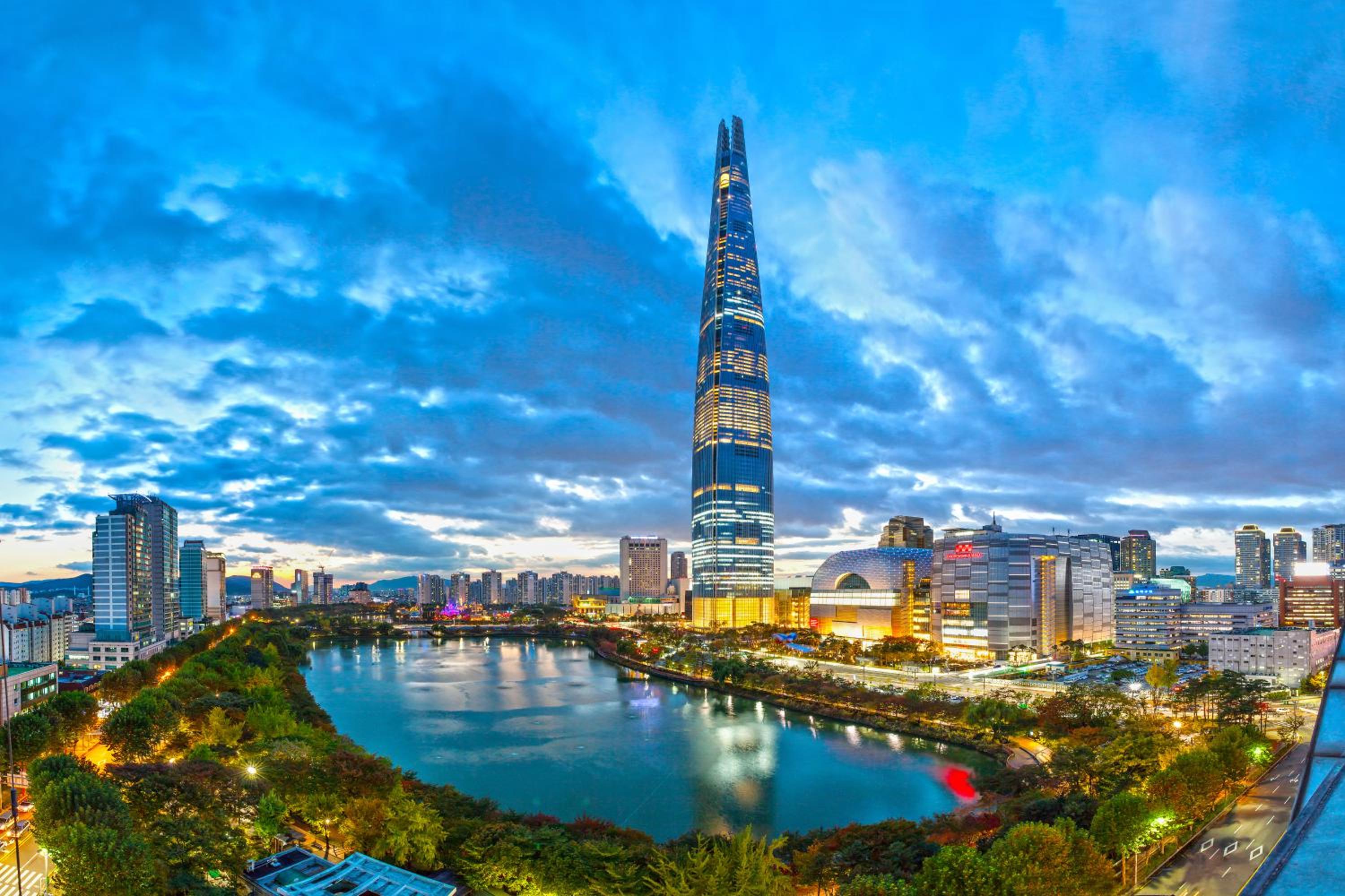 Lotte World Tower pierces Seoul's twilight sky, its illuminated facade reflecting across the Han River while the city's glittering metropolis spreads toward distant mountains.