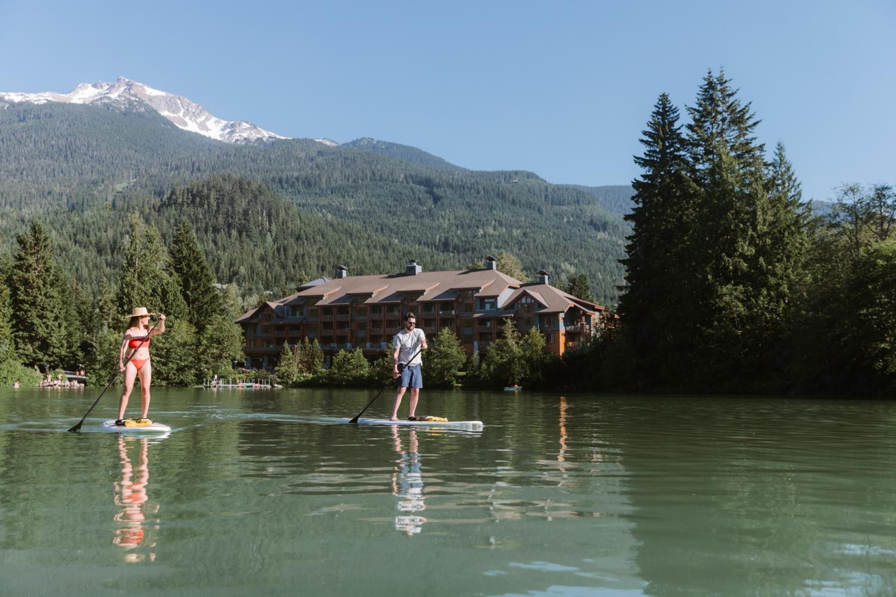 Paddleboarders glide across pristine alpine waters while Whistler's snow-capped peaks frame the lodge's timber architecture—mountain serenity at its most authentic.