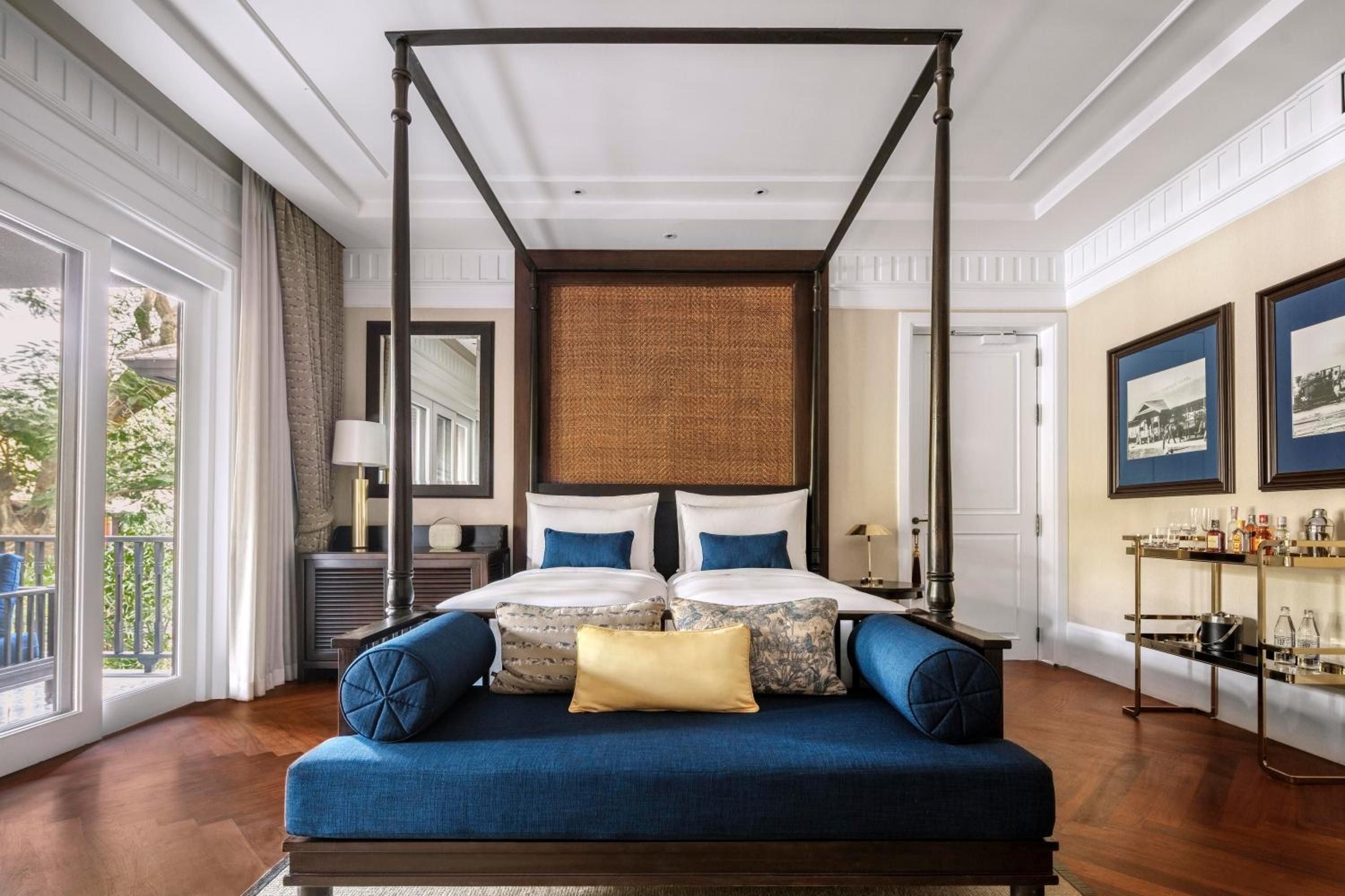 Woven rattan headboards and rich navy velvet create an elegant colonial-meets-contemporary aesthetic in this spacious suite, where polished teak floors catch the tropical light streaming through French doors.