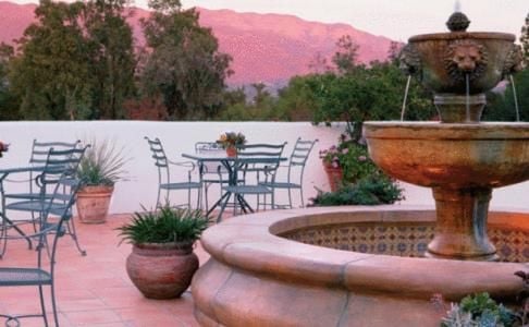 Pink-hued mountains frame an intimate courtyard where wrought-iron chairs invite sunset conversations beside a bubbling stone fountain and terra cotta planters.