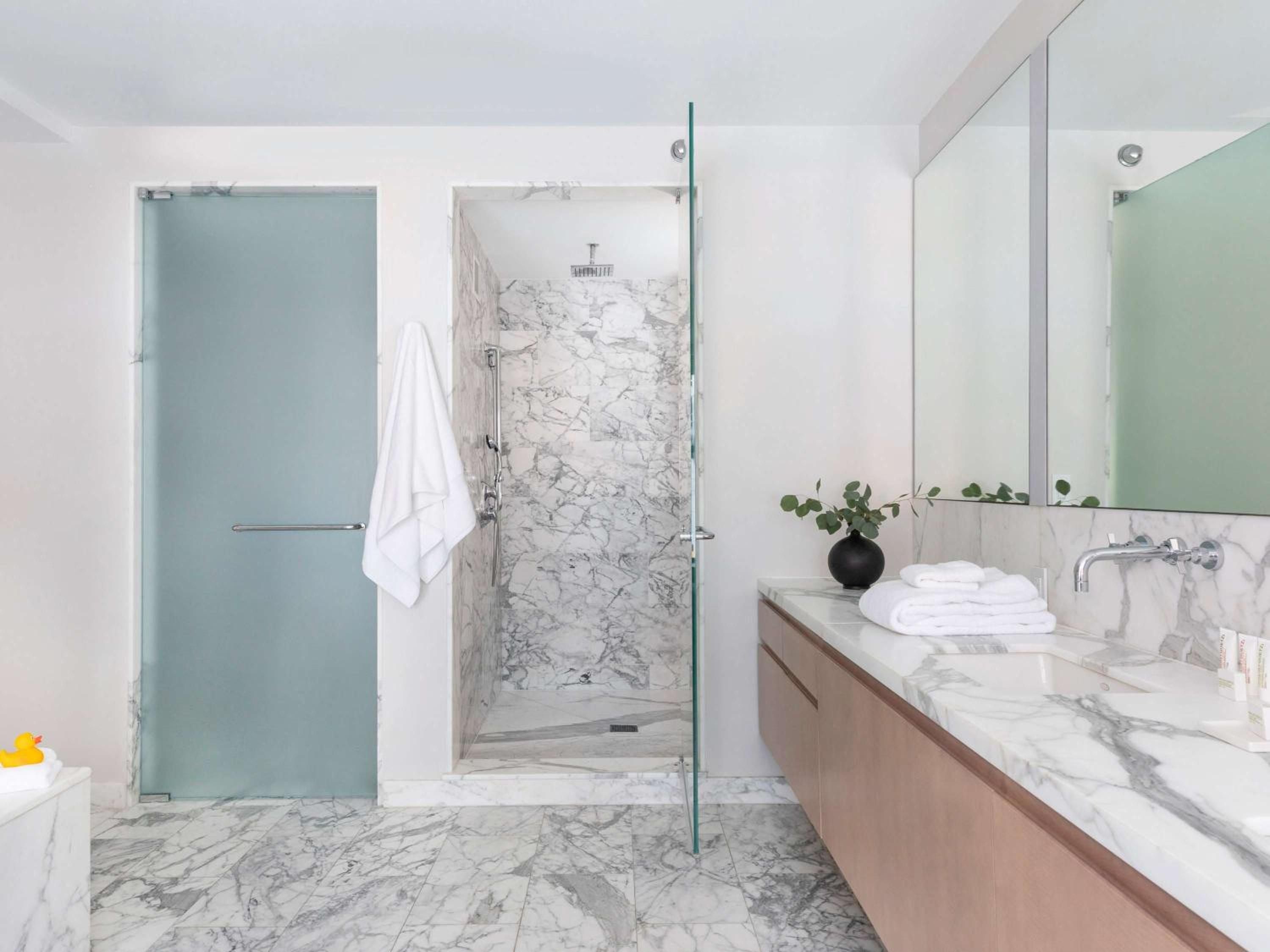 Calacatta marble cascades across floors and walls while warm wood vanities create intimate luxury in this serene spa-like retreat with glass-enclosed rainfall shower.