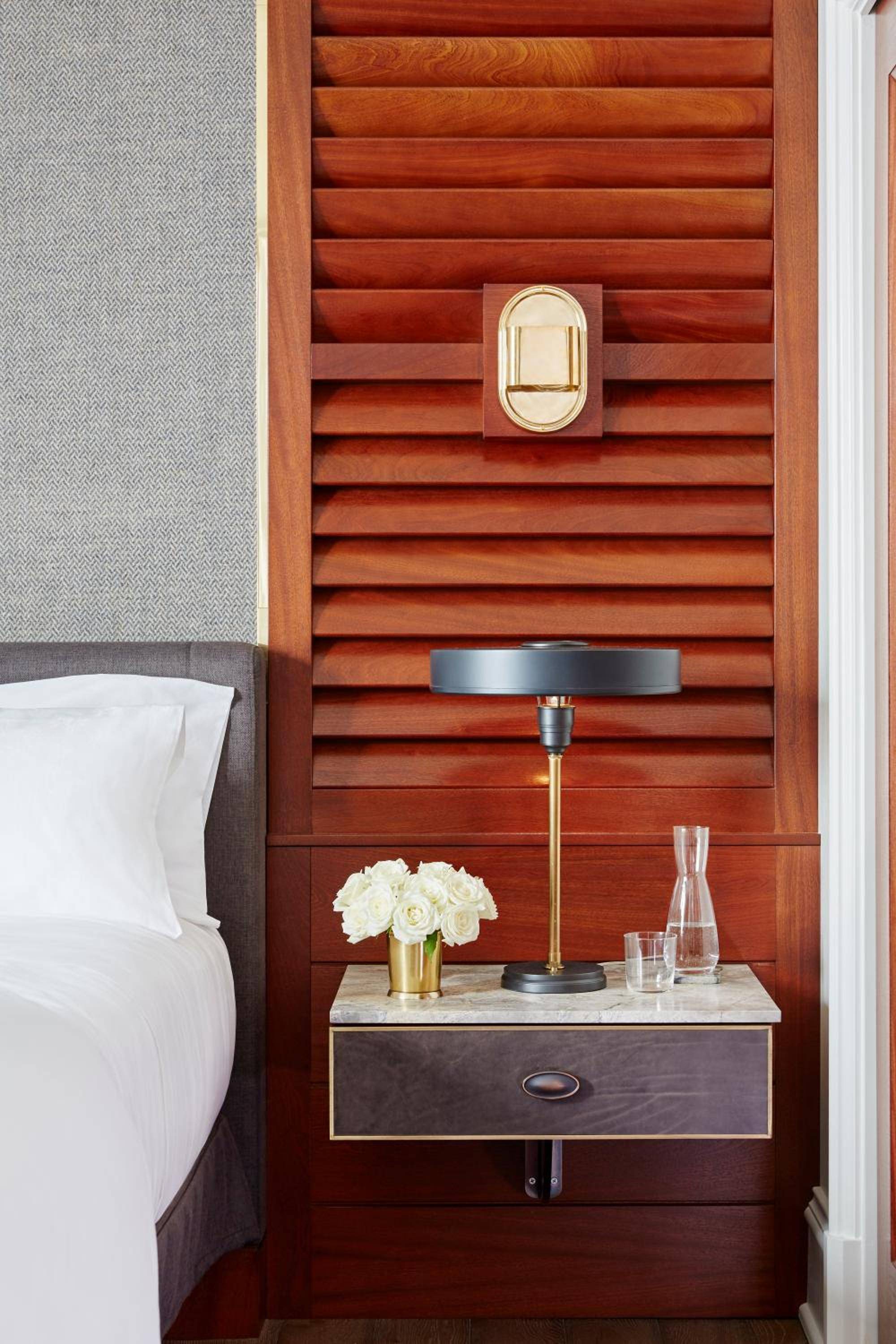 Warm mahogany shutters frame a floating nightstand topped with brass accents and fresh white roses, creating an intimate maritime elegance in guest quarters.