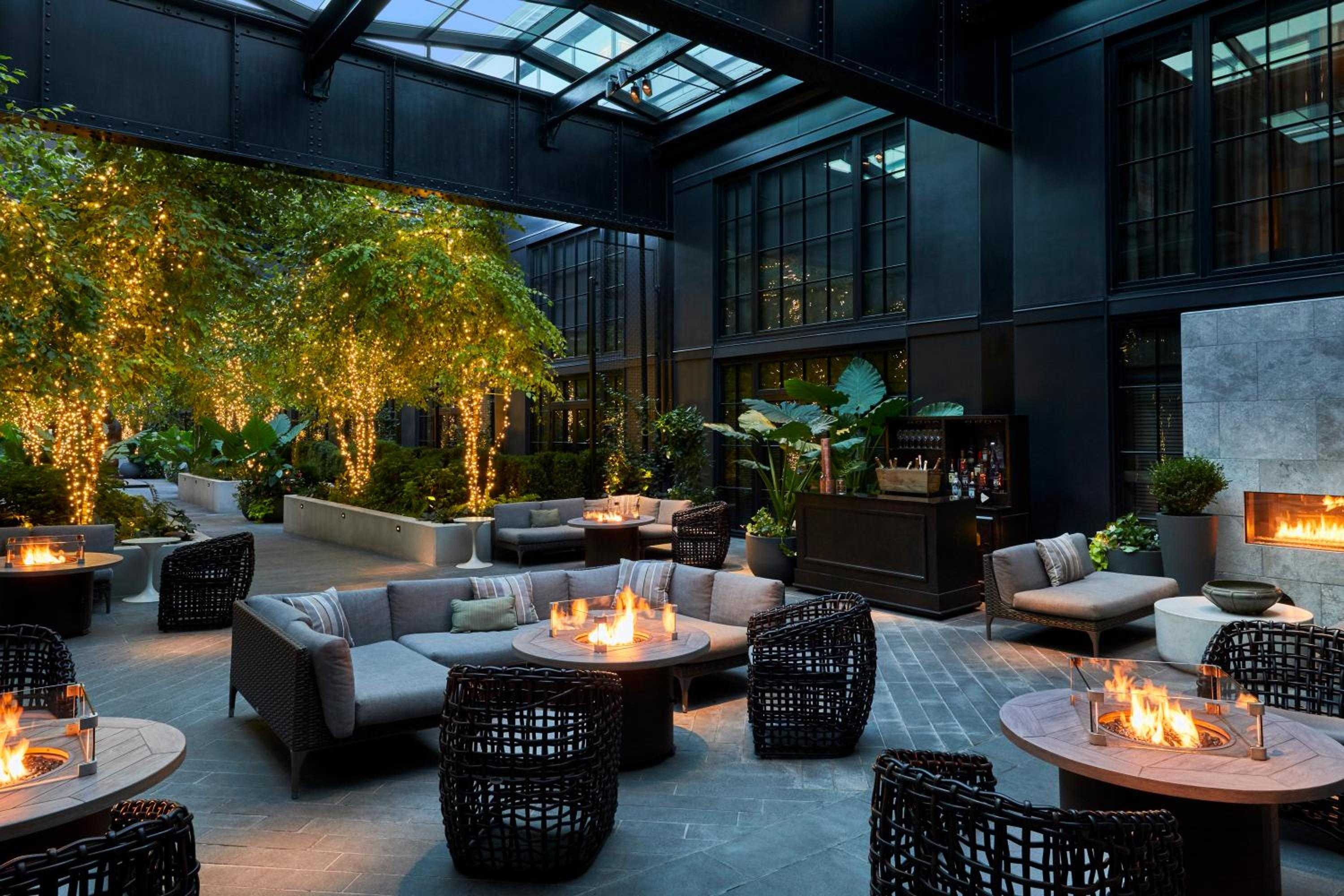 String lights cascade through leafy branches above intimate fire tables, where guests gather beneath Baltimore's skyline in this enchanting covered courtyard sanctuary.