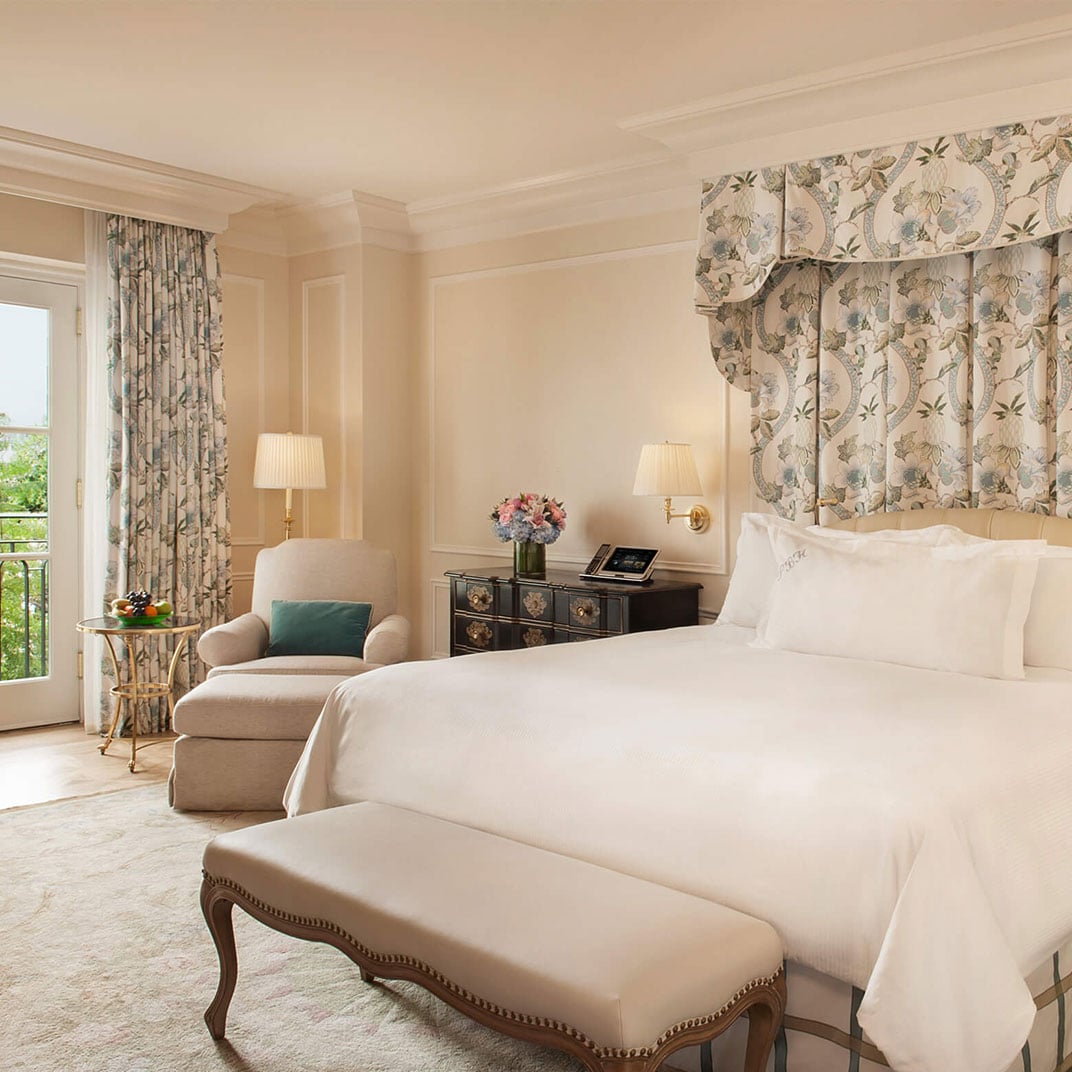 Soft afternoon light filters through botanical curtains onto crisp white linens, while an elegant upholstered headboard and French doors create sanctuary-like tranquility in this refined guest room.