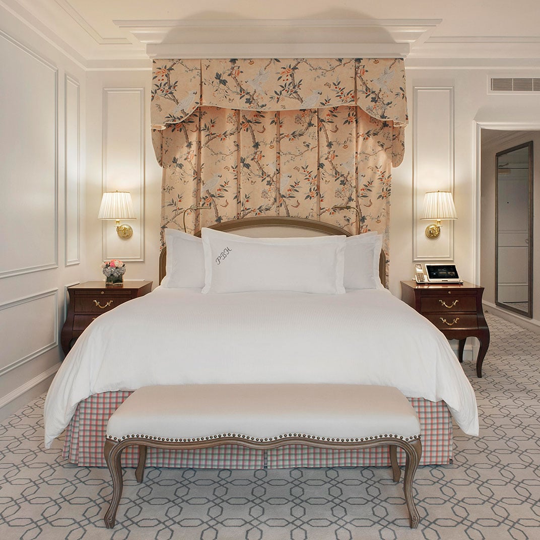 Soft morning light illuminates crisp white linens and floral chinoiserie headboard, while brass sconces cast warm pools of light across detailed crown molding and geometric carpeting.