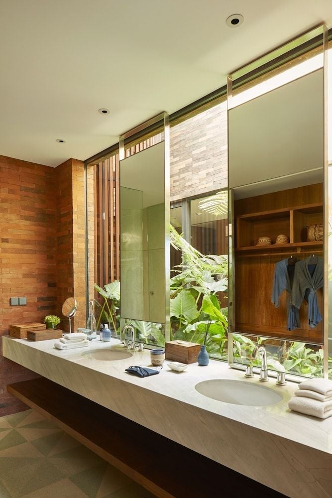 Natural stone and warm wood create a spa-like sanctuary where floor-to-ceiling windows frame lush tropical greenery, blurring boundaries between indoor luxury and Bali's verdant landscape.