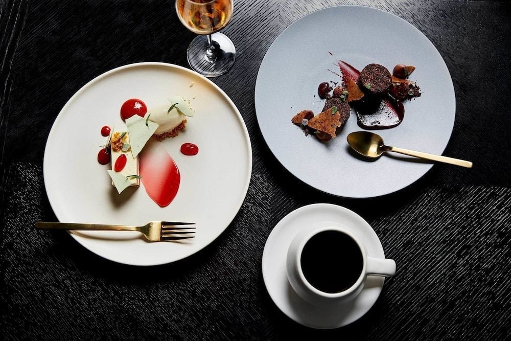Artfully plated desserts and rich coffee create an intimate finale to dinner, with golden spoons catching light against charcoal-dark wood grain.