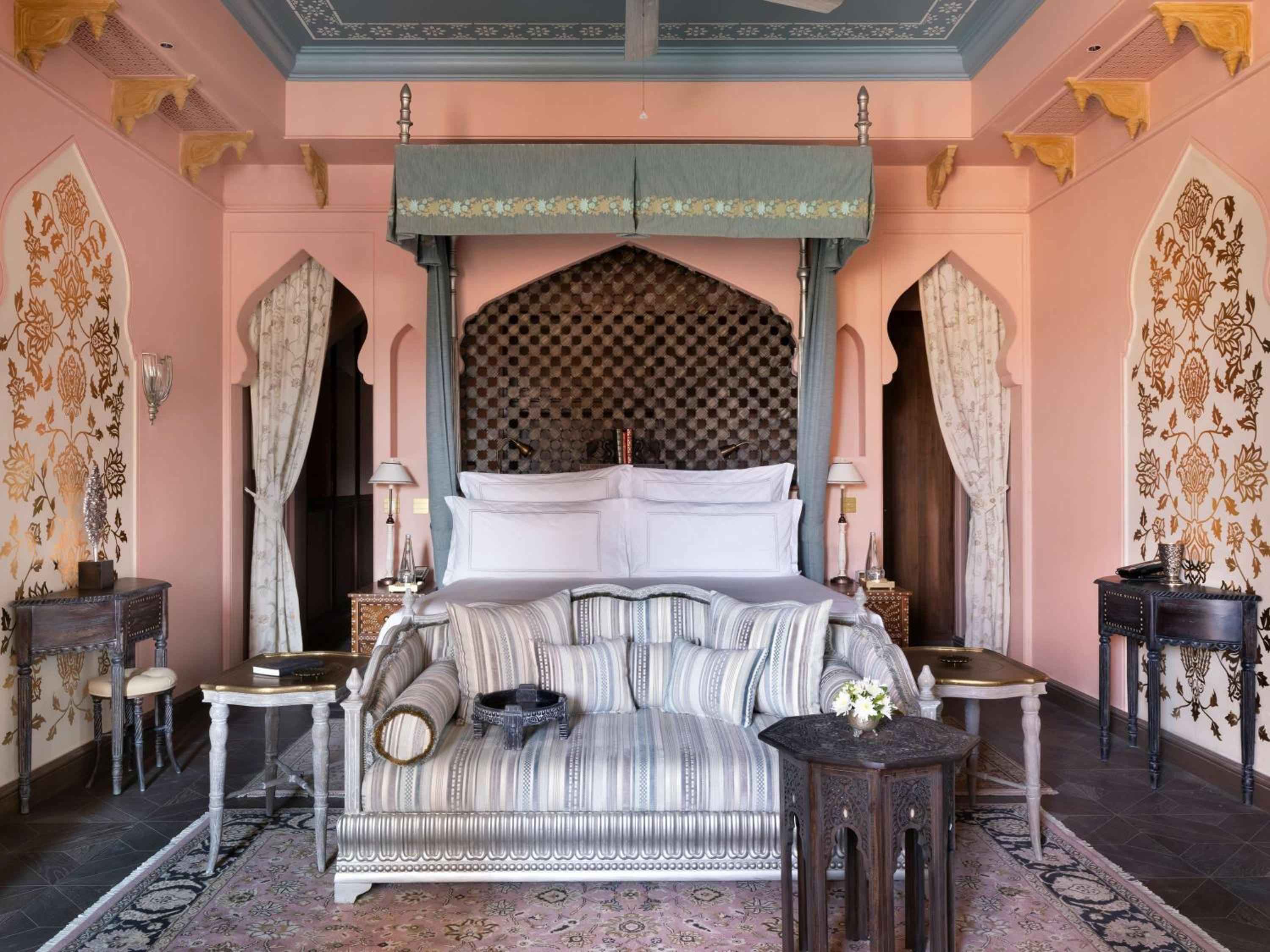 Moorish arches frame a canopied bed beneath hand-painted ceilings, where rose-hued walls and intricate tilework create an enchanting maharaja's retreat.