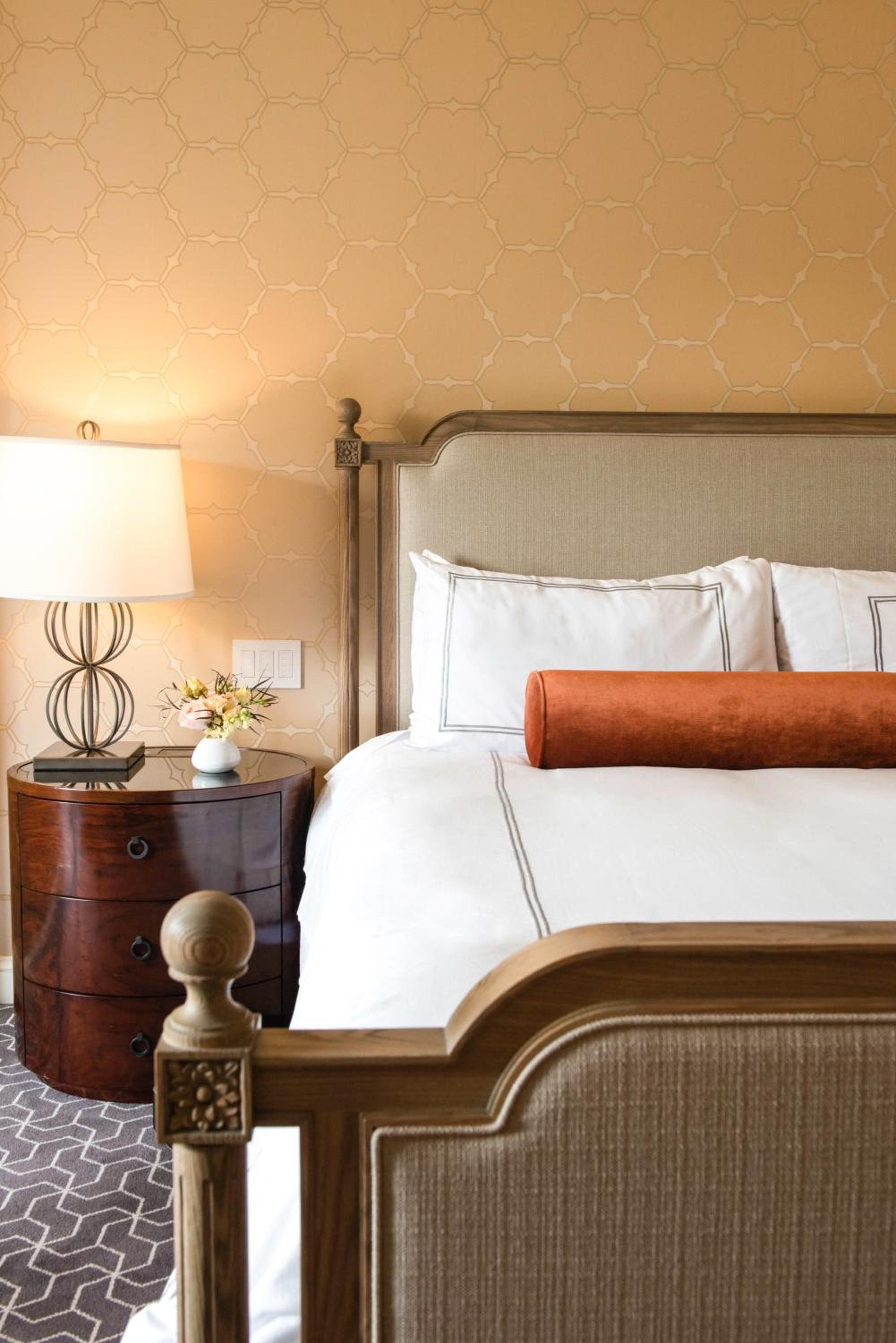 Honeycomb wallpaper casts warm amber light across crisp white linens and burnished wood, while an orange bolster adds a perfect pop of color to this sophisticated sanctuary.