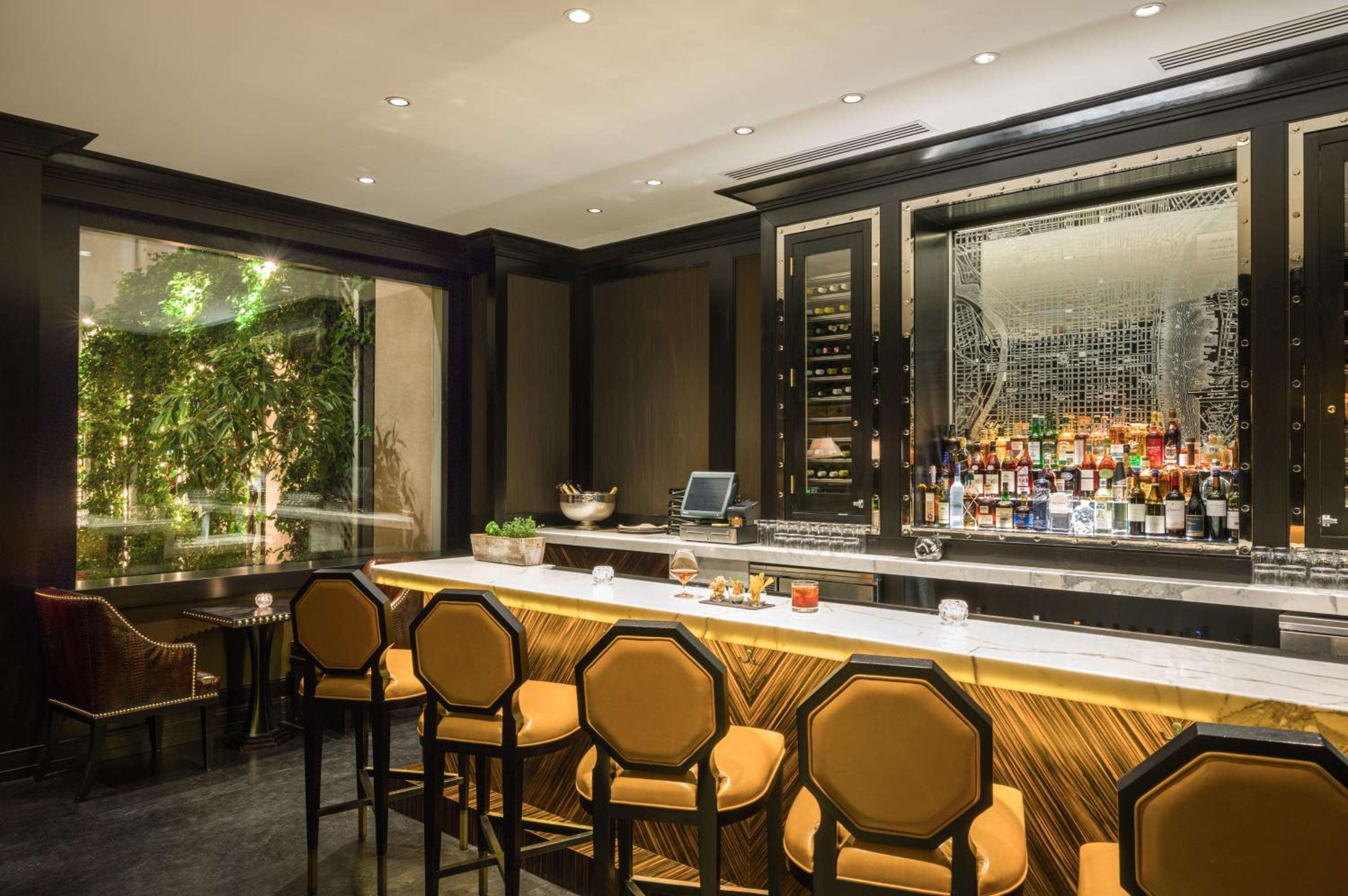 Honeyed leather barstools line the marble counter where premium spirits catch ambient light, while verdant garden views soften the sophisticated wood-paneled setting.