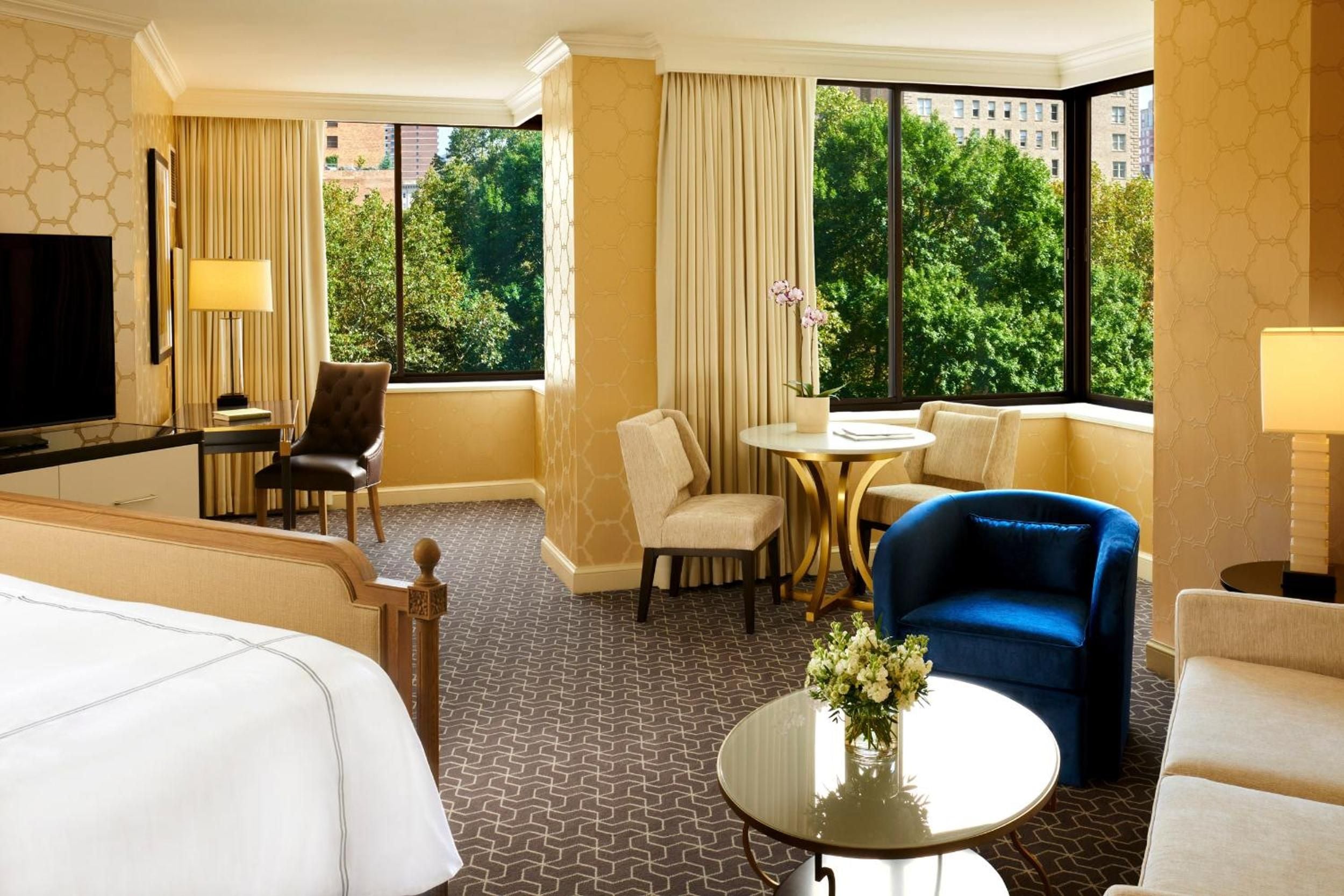 Sunlight streams through floor-to-ceiling windows of this elegant suite, where a sapphire velvet chair anchors the sophisticated seating area overlooking leafy Rittenhouse Square.
