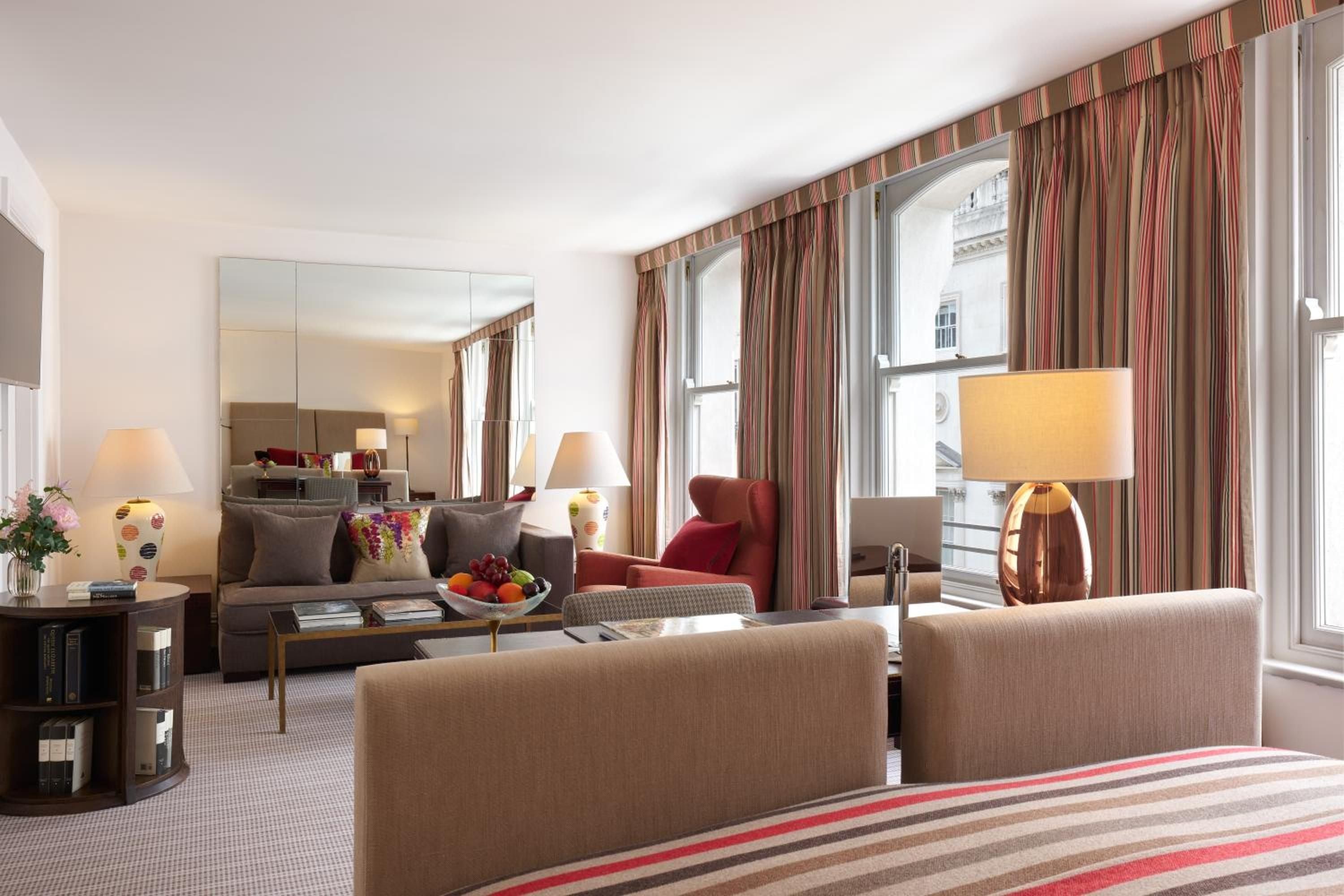 Plush taupe armchairs and striped curtains create an elegant sitting area where afternoon light filters through tall windows, revealing glimpses of Mayfair beyond.