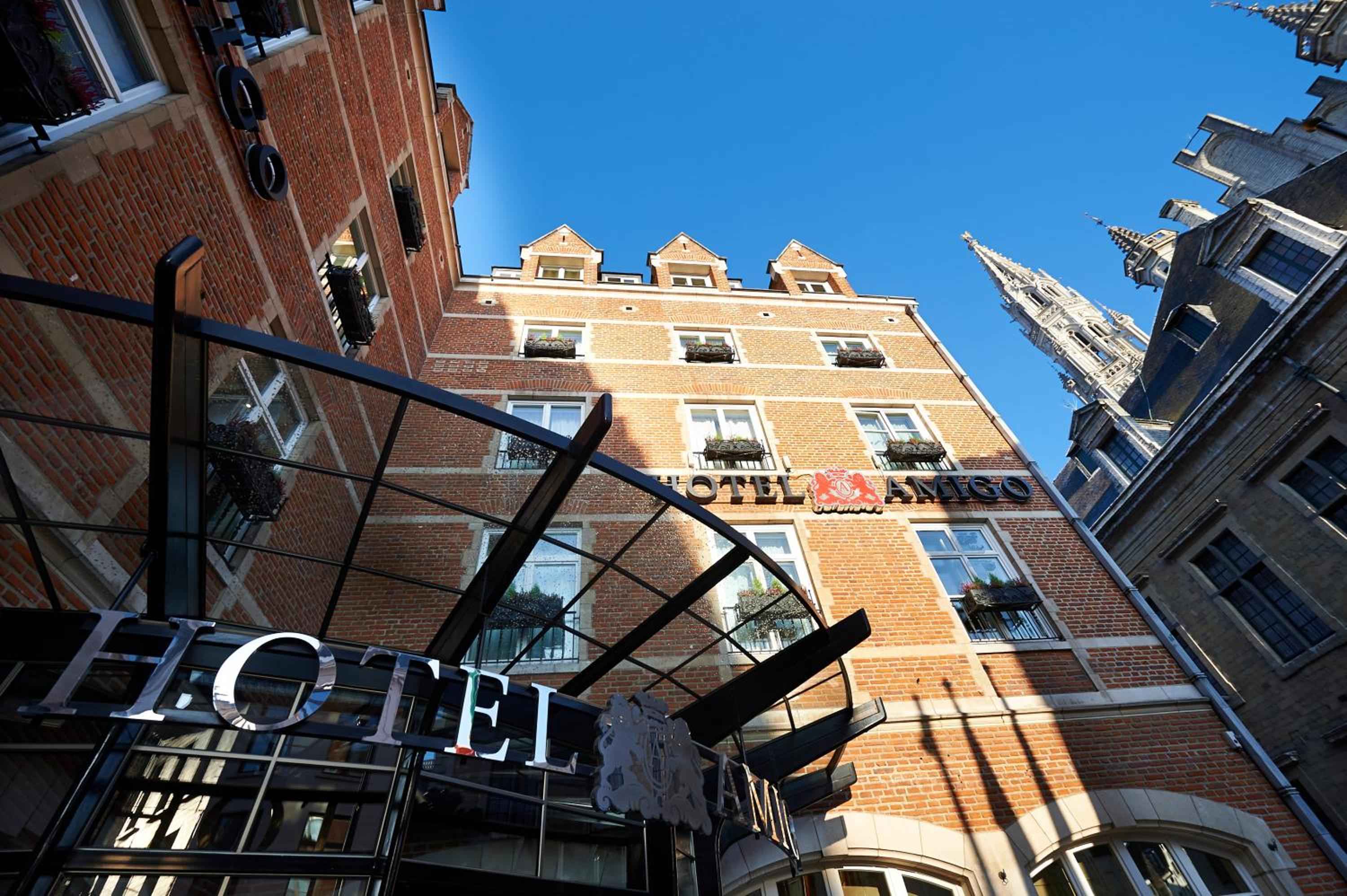 Warm brick facades and elegant ironwork frame Hotel Amigo's welcoming entrance, where Brussels' historic charm meets refined hospitality beneath azure skies.